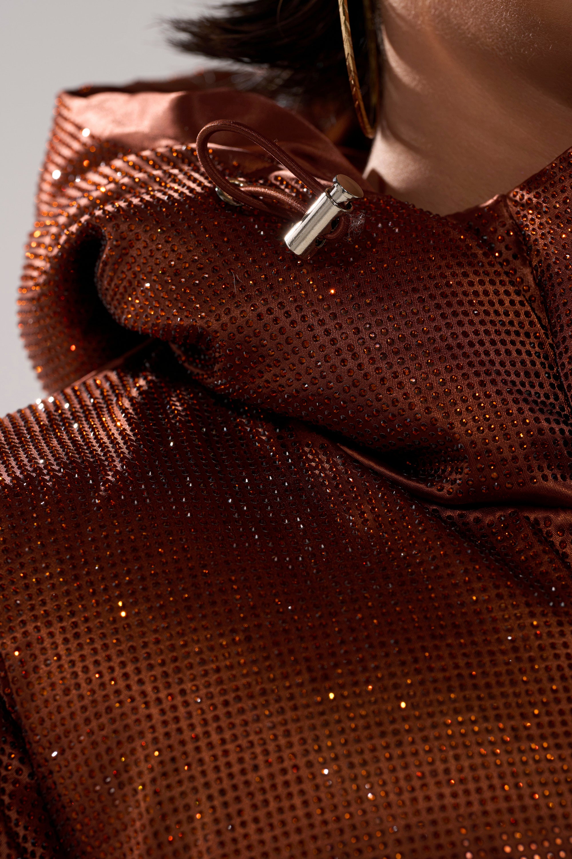 Close-up of a person wearing the FAYE RHINESTONE PUFFER IN COPPER, featuring sparkling copper sequins and metal-tipped drawstrings. Part of their jaw and a hoop earring can be seen, highlighting the puffer's luxe shine.