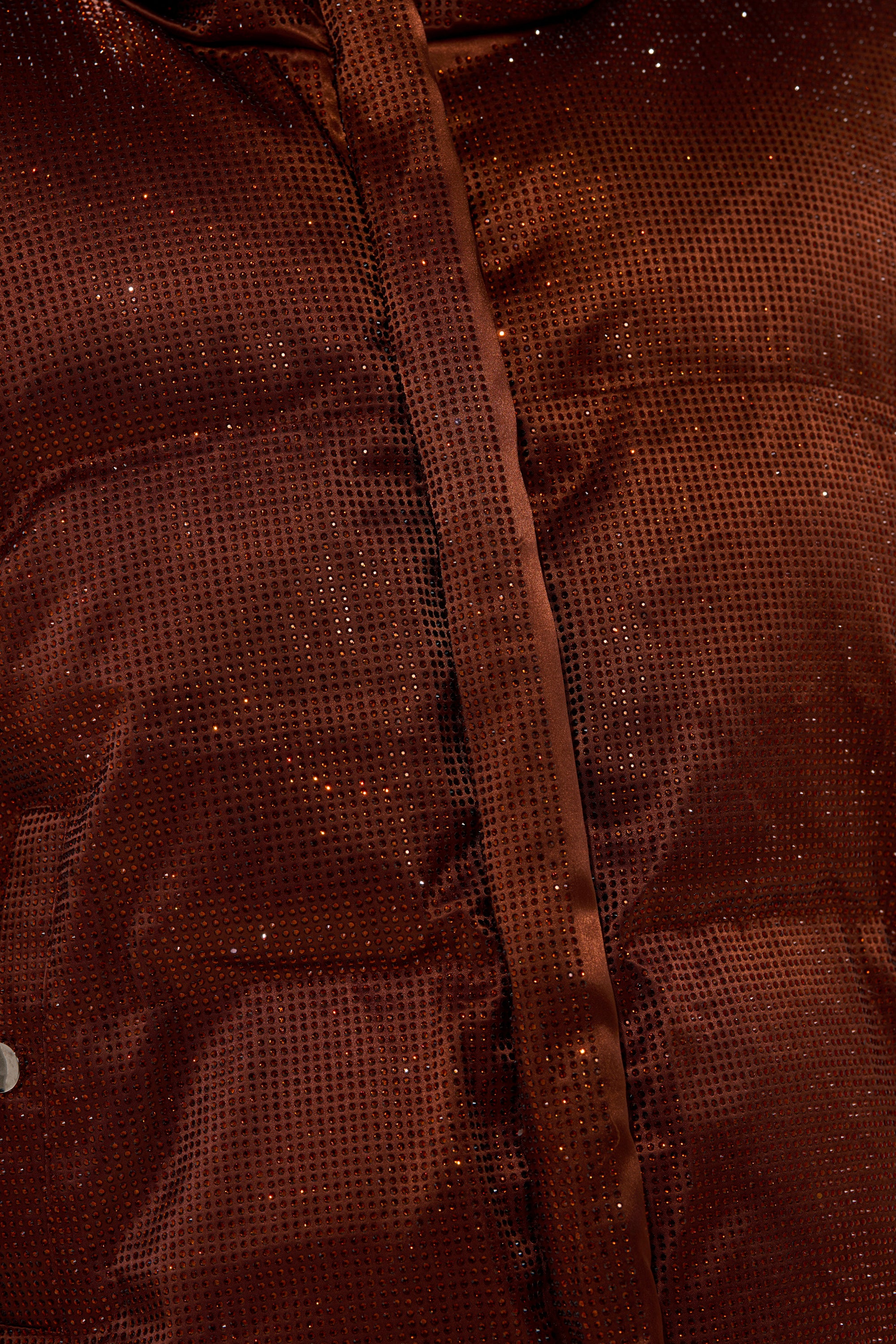 Close-up of shiny brown fabric with sequins and a vertical seam, resembling the glossy finish of the FAYE RHINESTONE PUFFER IN COPPER, suggesting it’s part of this stylish outerwear piece.