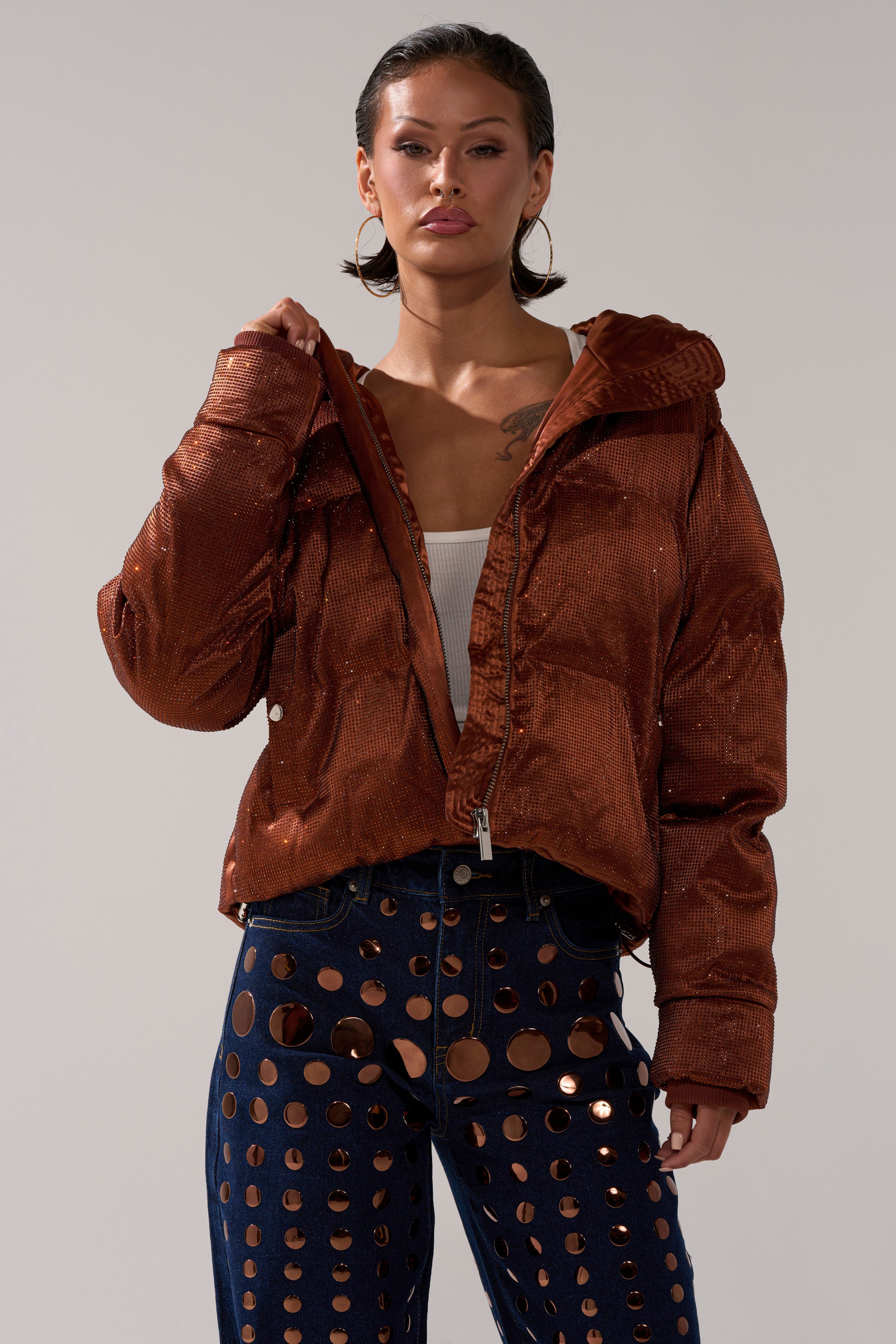 A woman stands confidently against a plain background, wearing the FAYE RHINESTONE PUFFER IN COPPER over a white crop top and navy blue pants with large circular cutouts.