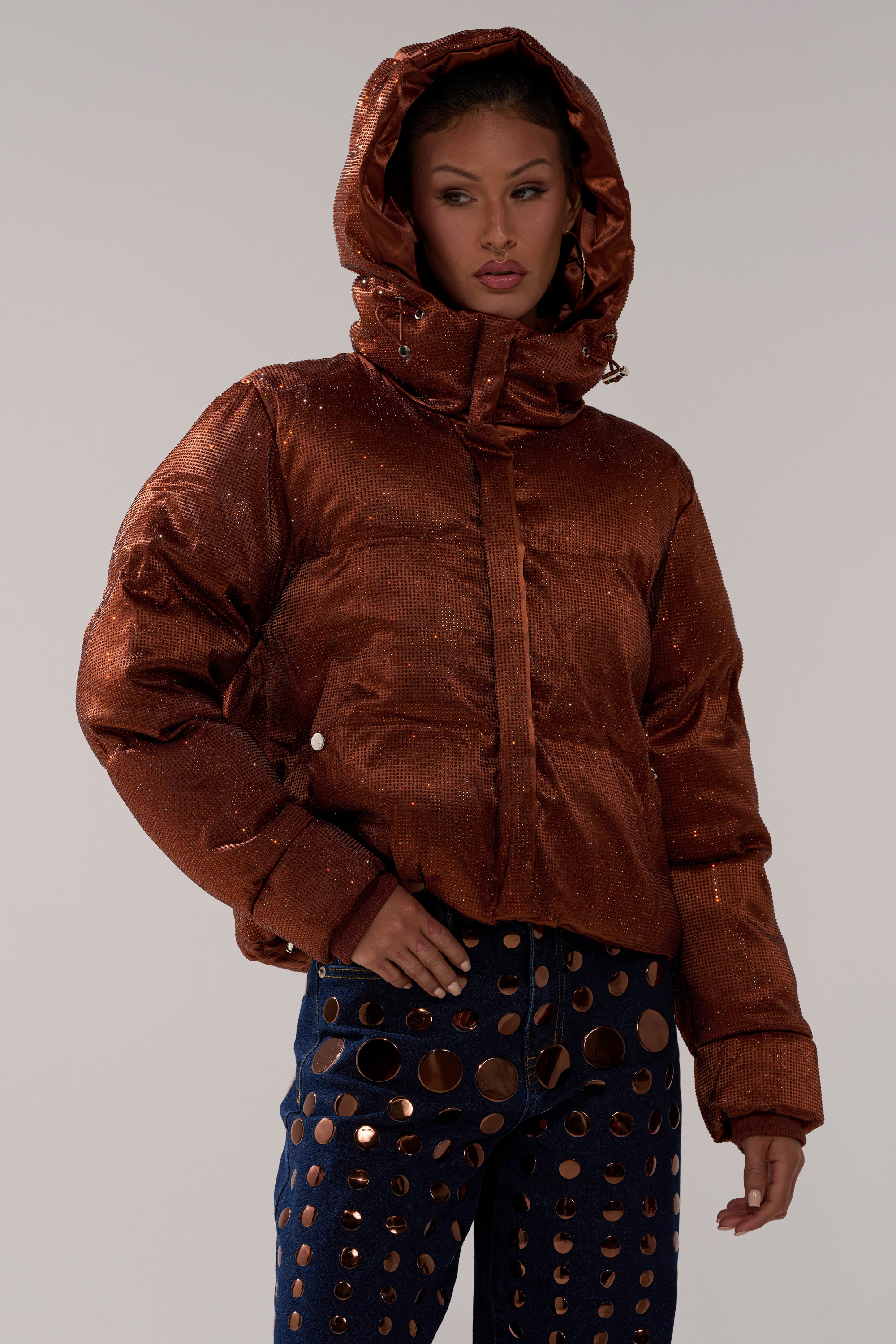 A woman poses against a plain light background wearing the FAYE RHINESTONE PUFFER IN COPPER and navy blue pants with large metallic circular cutouts.