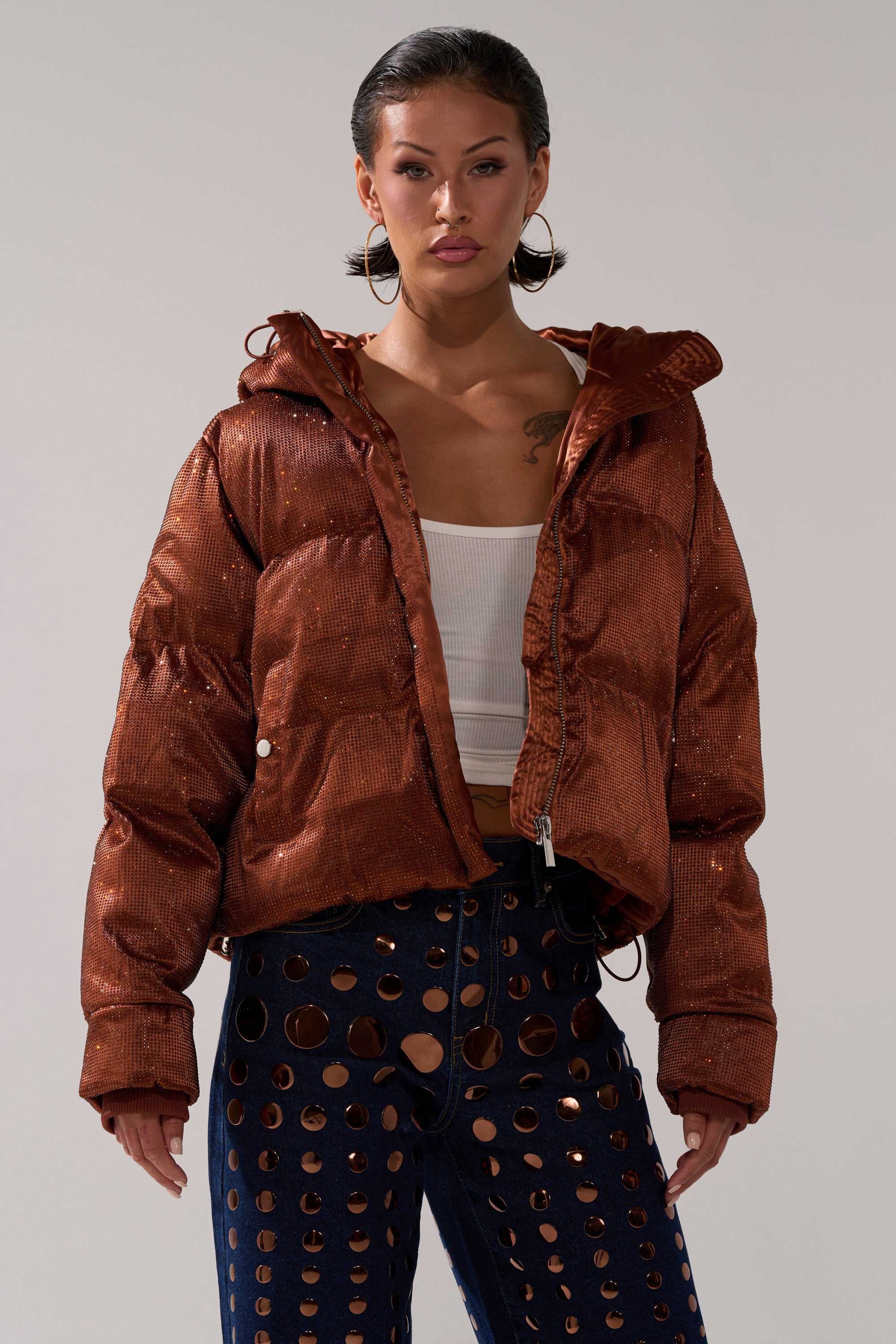 Wearing the FAYE RHINESTONE PUFFER IN COPPER over a white crop top and navy pants with large circular cutouts, the person stands against a plain background.