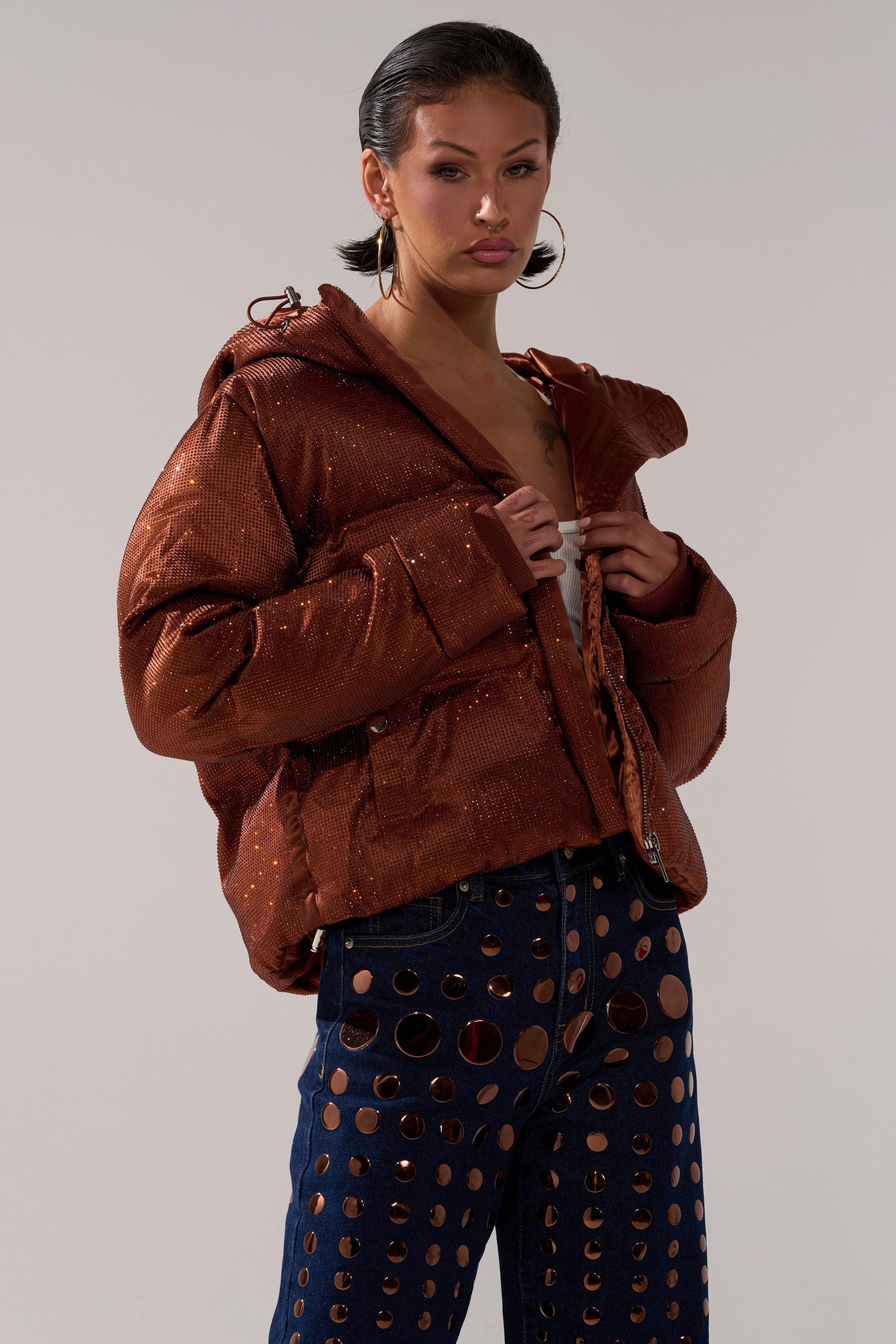 A person wearing the FAYE RHINESTONE PUFFER IN COPPER and dark blue pants with large circular cutouts, posing against a plain gray background.