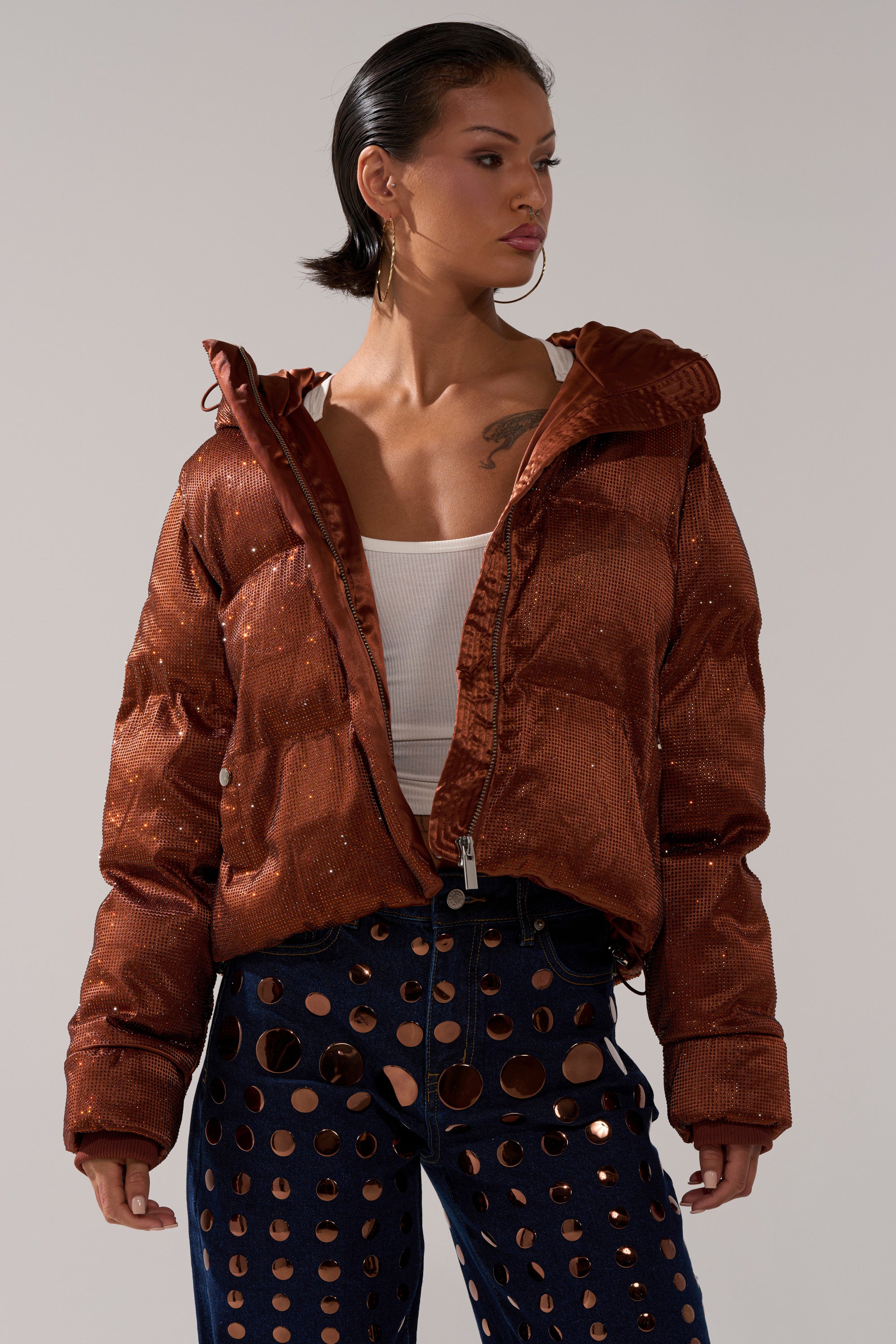 A person wears the FAYE RHINESTONE PUFFER IN COPPER over a white tank top and navy blue pants with large circular cutouts, standing against a plain background and looking confidently to the side.