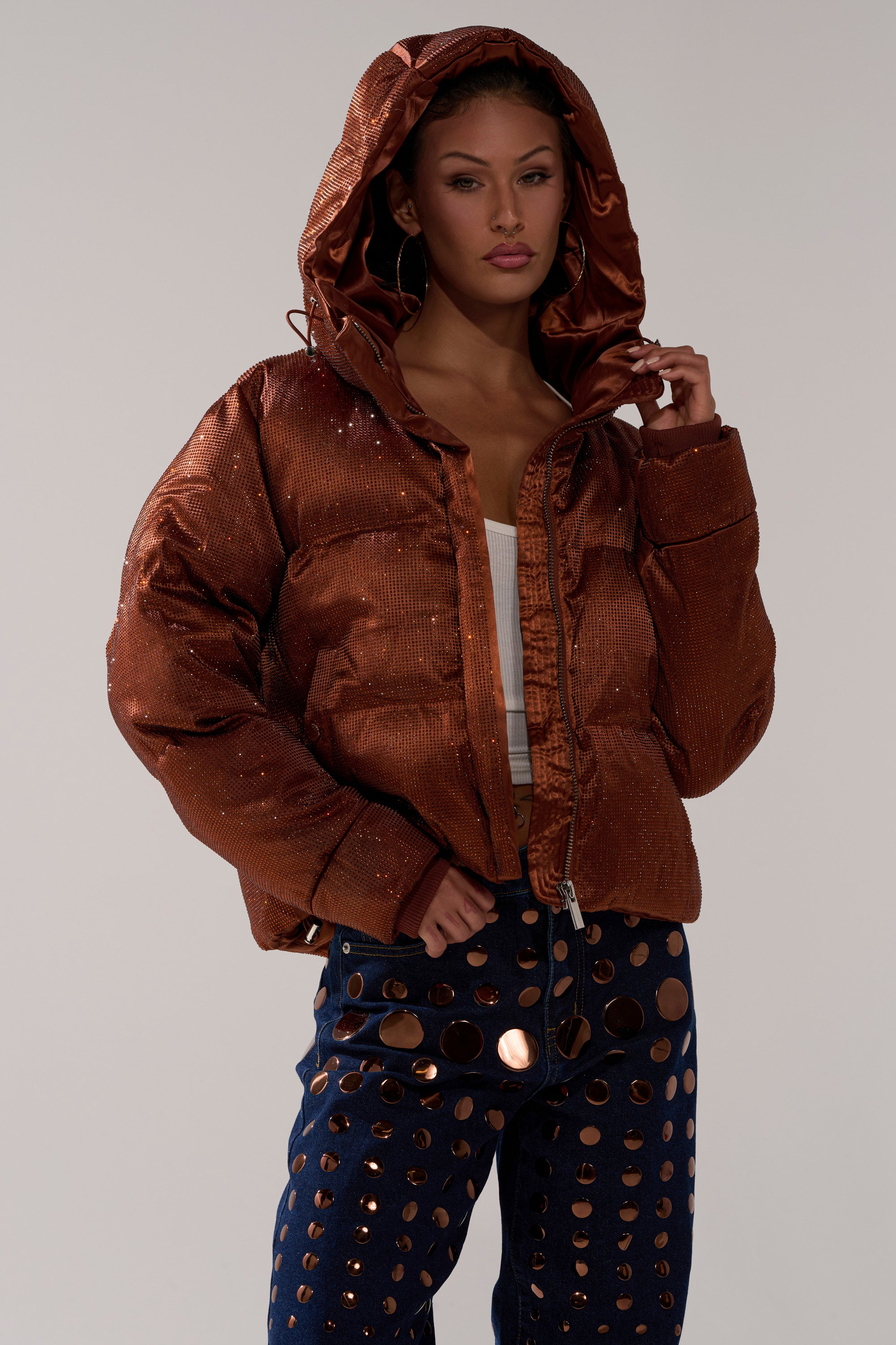 A woman poses confidently against a plain background in the FAYE RHINESTONE PUFFER IN COPPER, a shiny jacket styled with a white crop top and dark blue jeans featuring large metallic circular embellishments.