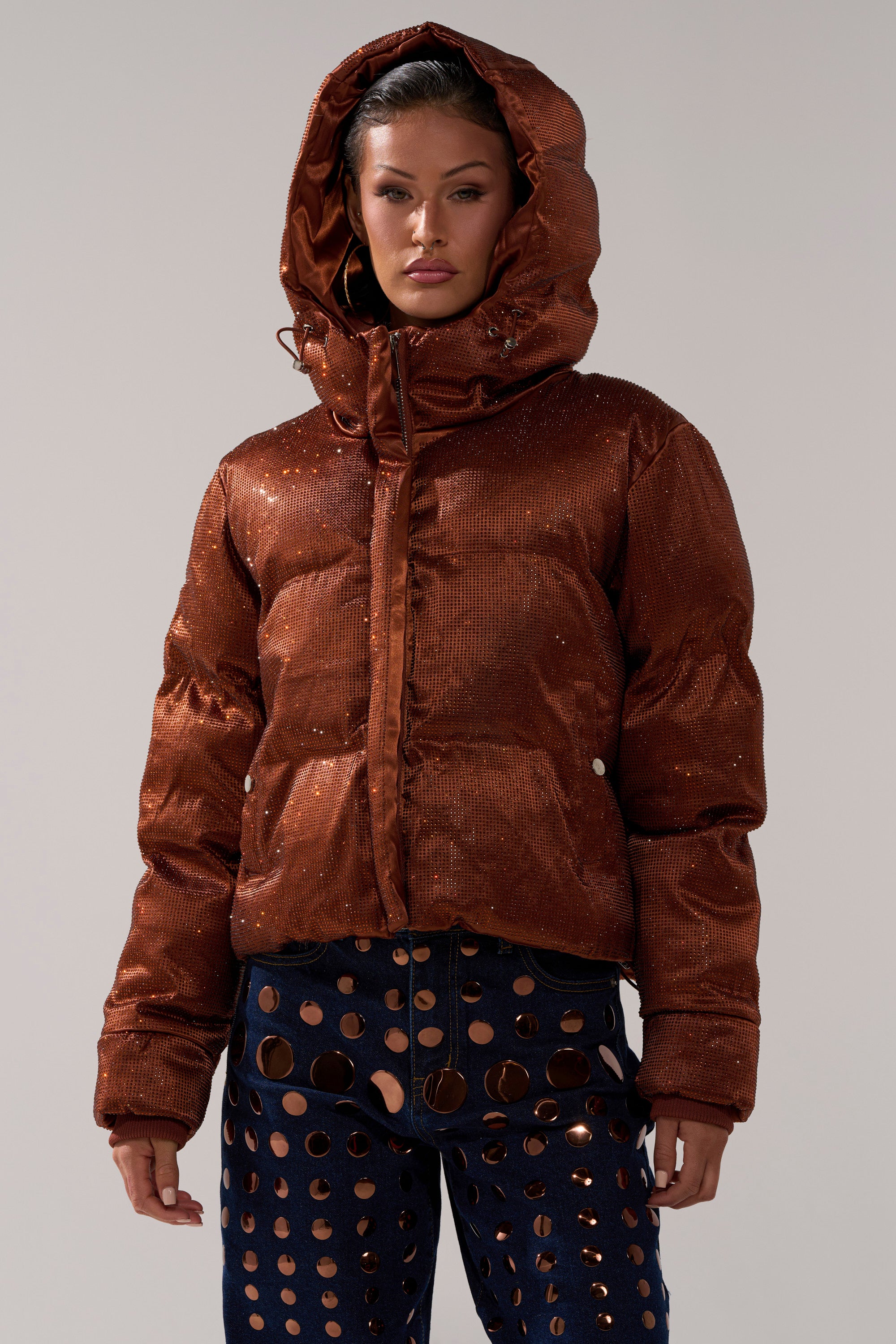 A woman wears the FAYE RHINESTONE PUFFER IN COPPER with a hood, paired with dark blue pants featuring large circular cutouts, standing against a plain background.