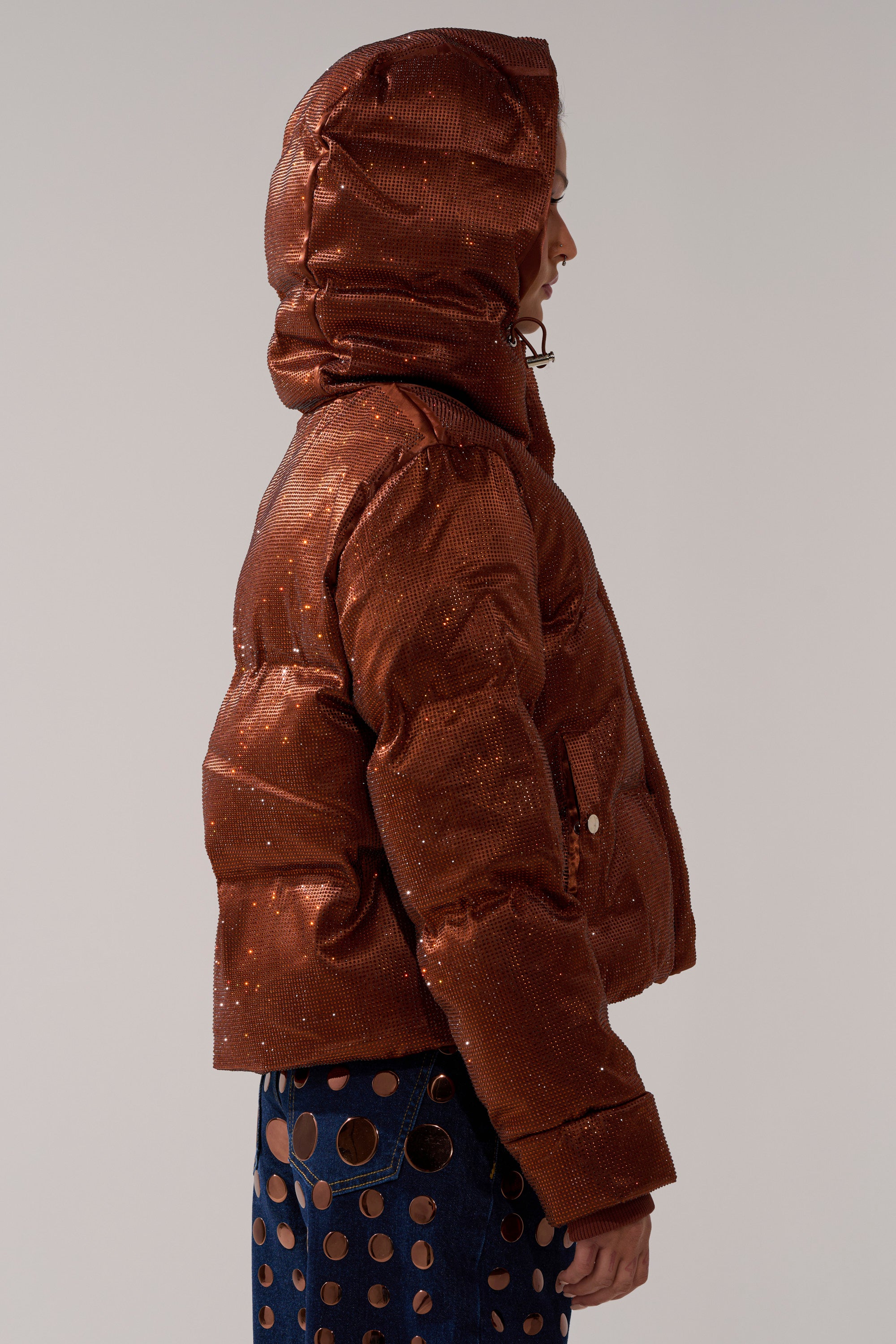 A person in profile wears the FAYE RHINESTONE PUFFER IN COPPER—a shiny, copper jacket with metallic accents—paired with dark blue pants featuring large circular cutouts. The background is plain and light-colored.
