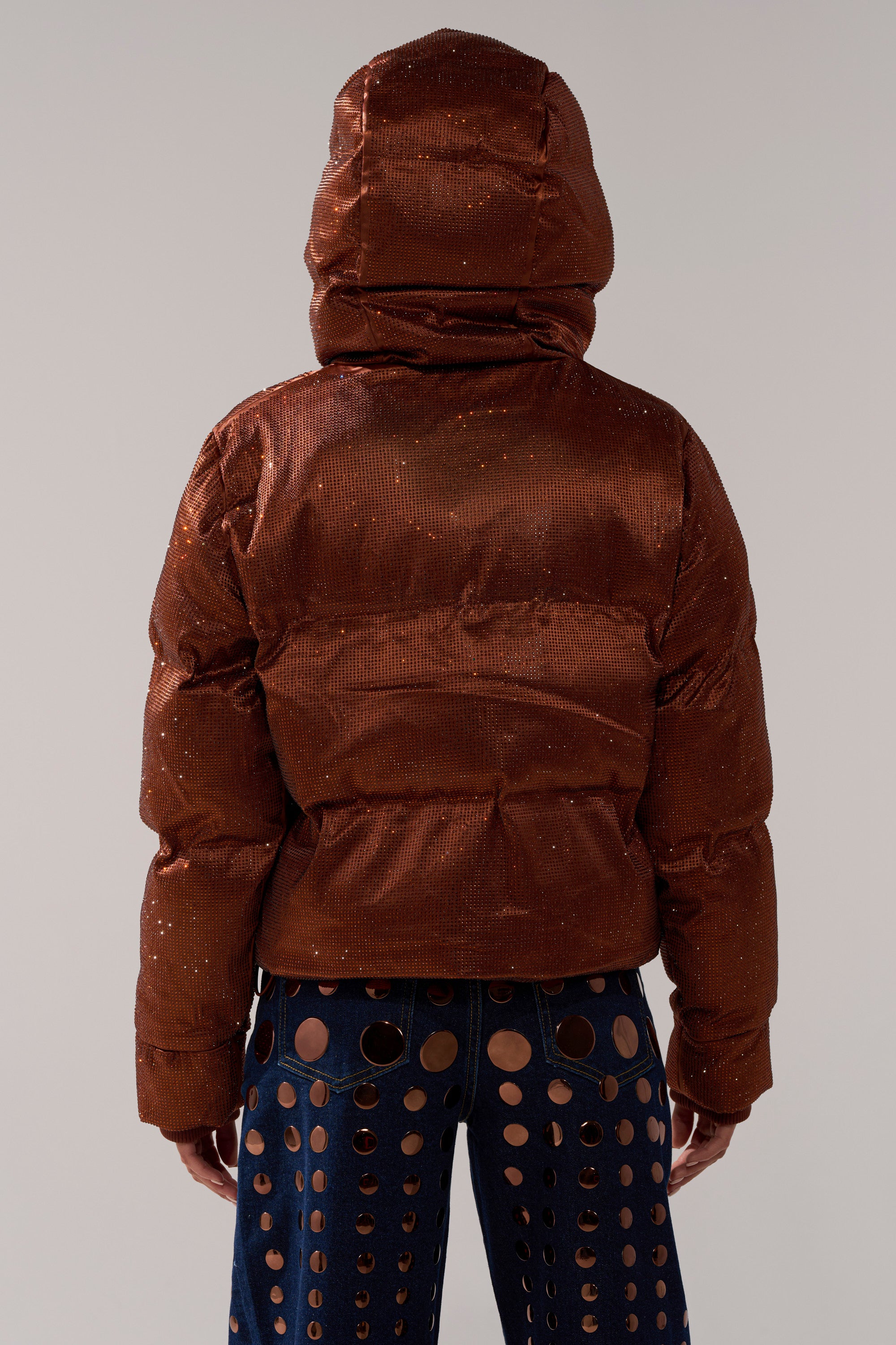 A person wears the FAYE RHINESTONE PUFFER IN COPPER, a sparkly brown hooded jacket, with blue high-waisted pants featuring large circular cutouts, shown from the back against a plain light background.