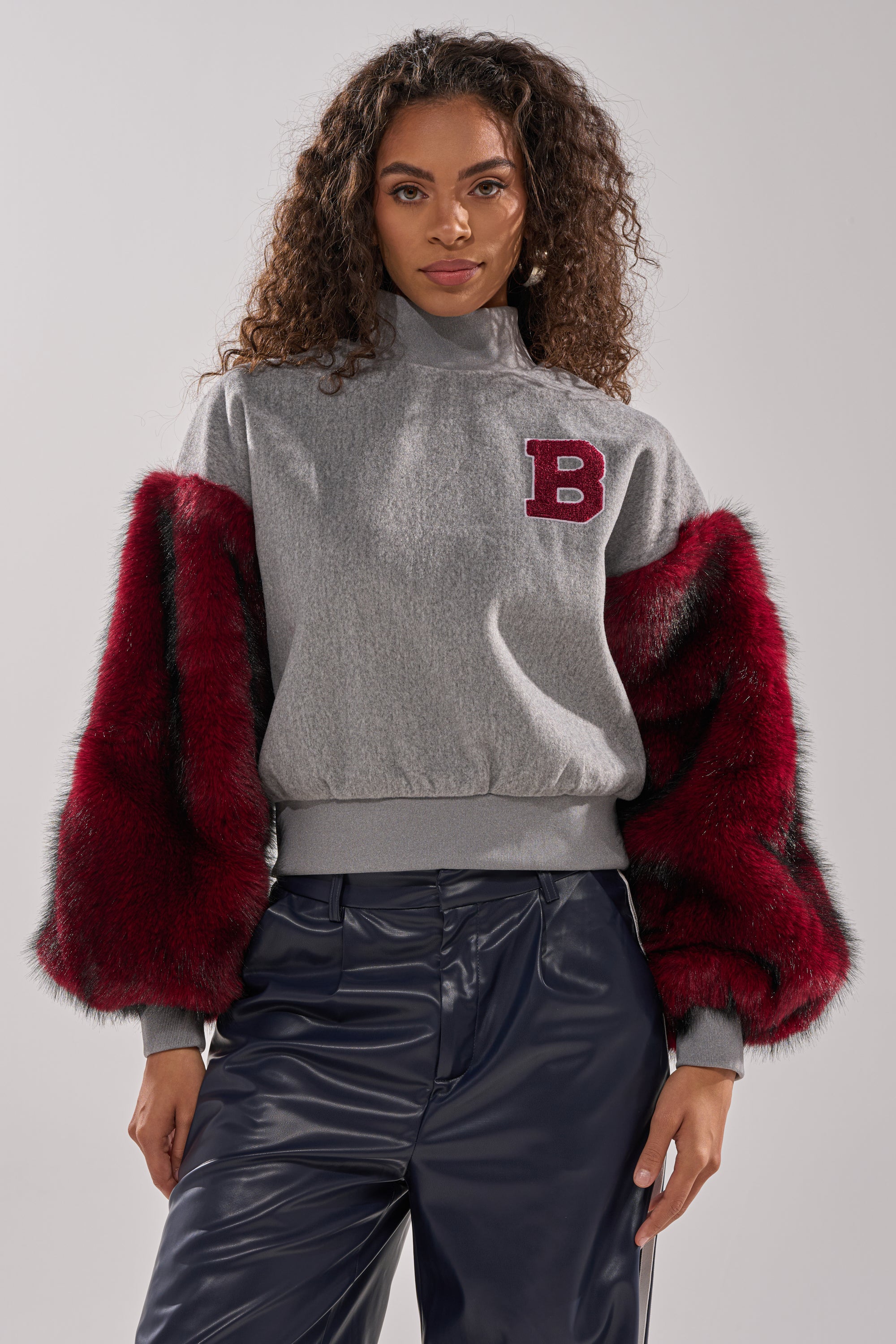 A woman with long curly hair wears the NEW EAST SIDE FAUX FUR SLEEVE SWEATSHIRT—gray with red "B" patch and bold red faux fur sleeves—styled with shiny navy blue pants, creating a striking streetwear look against a plain background.