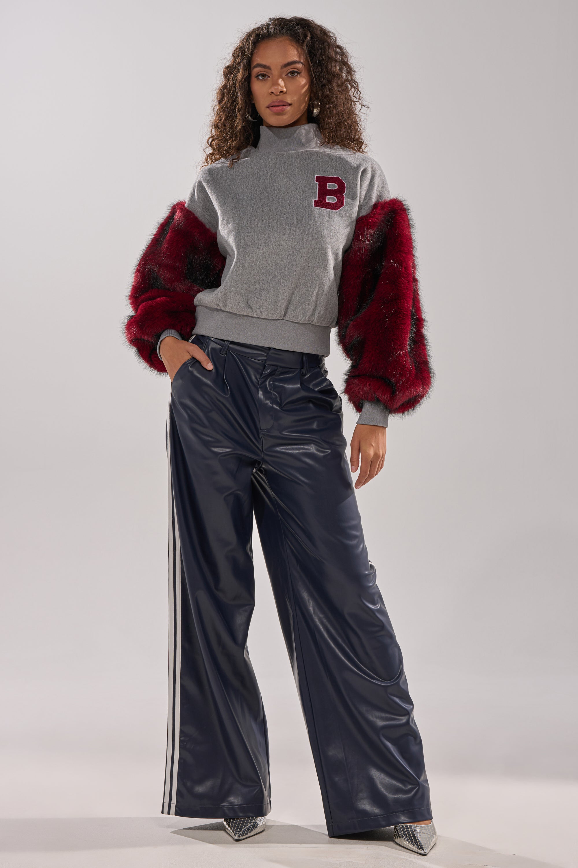 A woman stands confidently in the NEW EAST SIDE FAUX FUR SLEEVE SWEATSHIRT, featuring a letter "B" patch, styled with shiny navy wide-leg pants with white side stripes and silver pointed shoes.