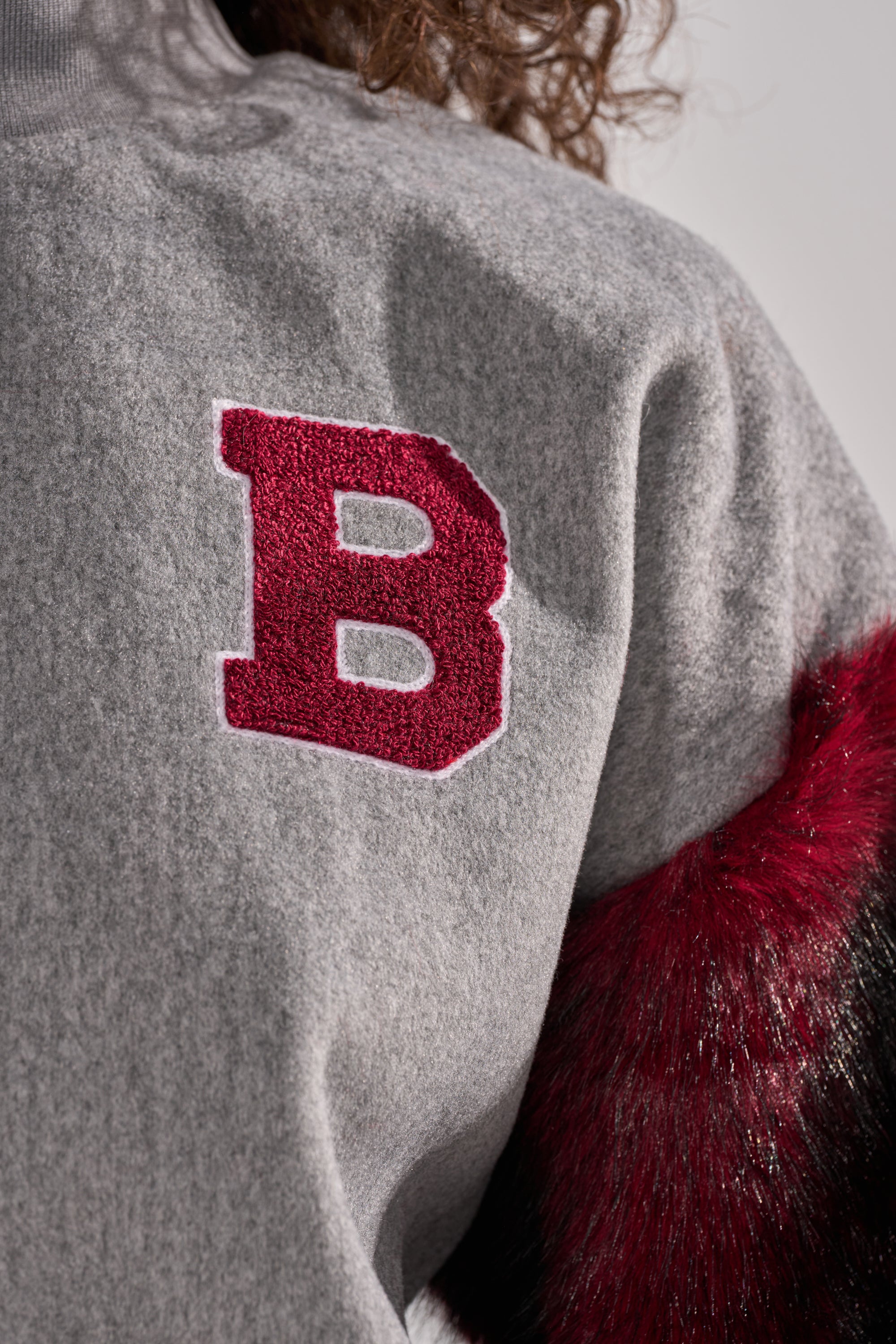 The NEW EAST SIDE FAUX FUR SLEEVE SWEATSHIRT features a gray body, bold red "B" chest patch, and red-black faux fur sleeve—perfect for standout streetwear style.