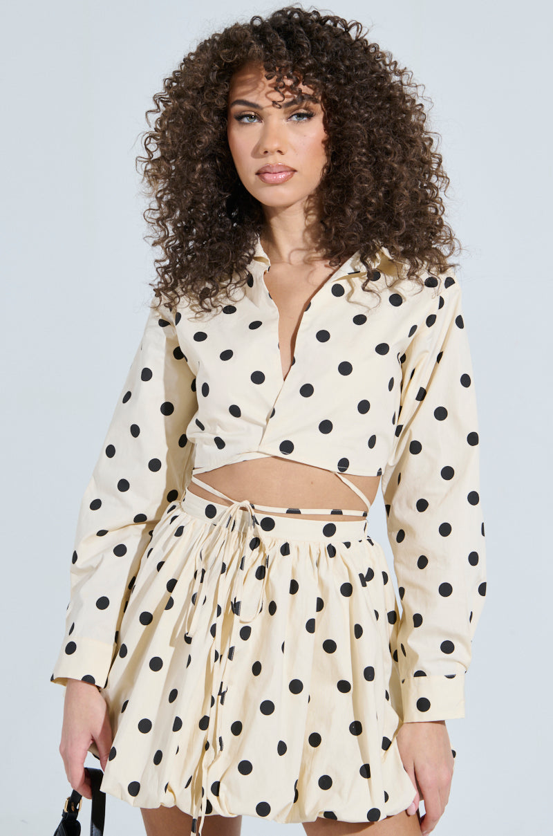 A woman with curly hair poses against a plain backdrop, donning the ALL ABOUT ME POLKA DOT POPLIN TOP in cream with a chic wrap-front design. The high-waisted skirt pairs seamlessly to complete the look.