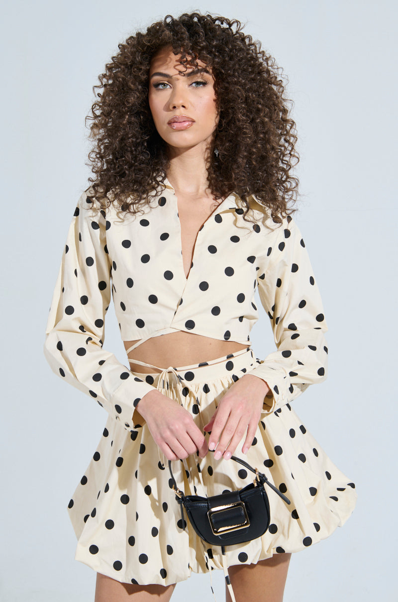 A woman with curly hair models the ALL ABOUT ME POLKA DOT POPLIN TOP in beige, paired with a wrap-front, drawstring skirt. She holds a small black handbag against a light background.