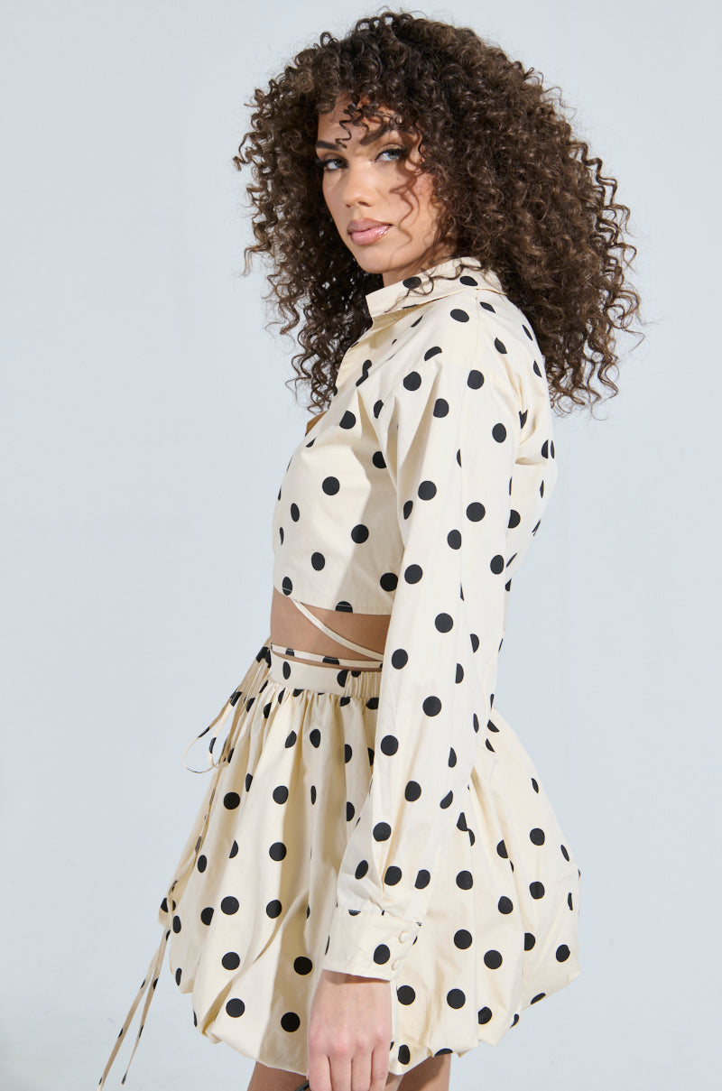 Against a light background, a person with curly hair exudes effortless charm in a creamy cotton poplin outfit from the "ALL ABOUT ME POLKA DOT POPLIN TOP," featuring black polka dots, paired with a short pleated skirt and string detailing as they glance over their shoulder.