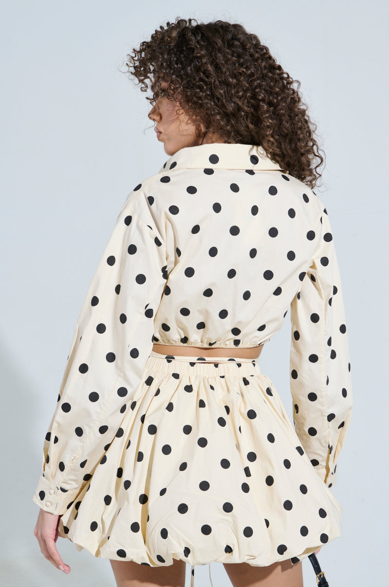 The curly-haired person wears the "ALL ABOUT ME POLKA DOT POPLIN TOP," featuring long sleeves and a wrap-front design in cream cotton poplin with black polka dots, as they stand back to the camera against a light background.