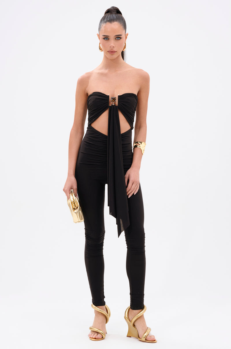 A woman poses confidently against a white background in the ONE OF A KIND CUTOUT JUMPSUIT—an elegant, strapless black jumpsuit in luxe stretch fabric—styled with gold heels, a gold bangle, a gold clutch, and a high ponytail.