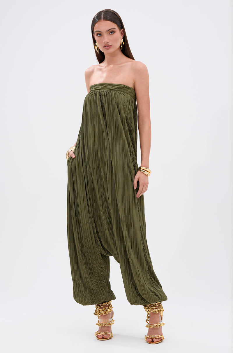 A woman models the FROM THE CITY DROP CROTCH JUMPSUIT—an olive, strapless, silky satin piece with wide harem legs. She pairs it with strappy gold heels and jewelry, standing against a simple light backdrop.