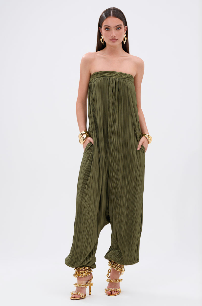 A woman wears the FROM THE CITY DROP CROTCH JUMPSUIT, a strapless, loose-fit olive satin piece with a pleated harem silhouette. She pairs it with gold hoop earrings, chunky bracelets, and chain-detailed heeled sandals against a plain white background.