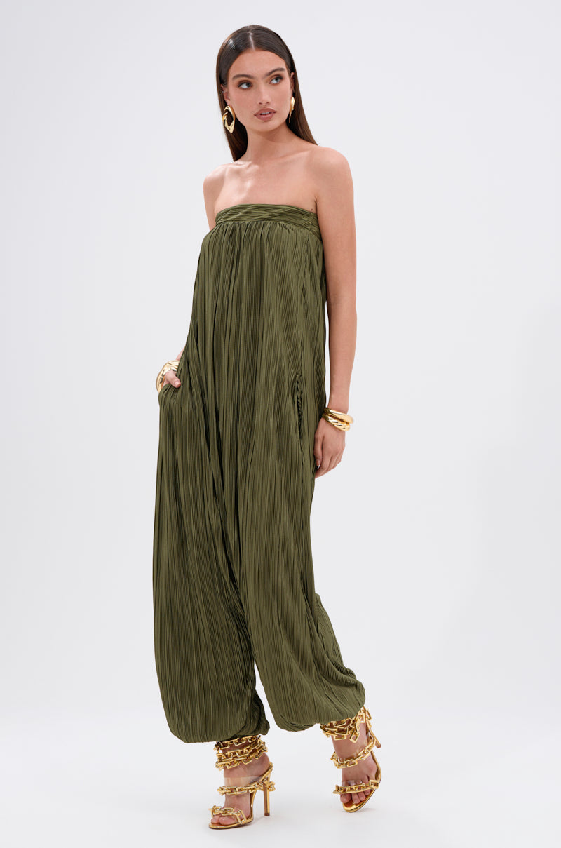 A woman stands against a white background wearing the FROM THE CITY DROP CROTCH JUMPSUIT, a strapless olive satin piece with wide harem-style legs gathered at the ankles, paired with gold hoop earrings, bangles, and high-heeled sandals.