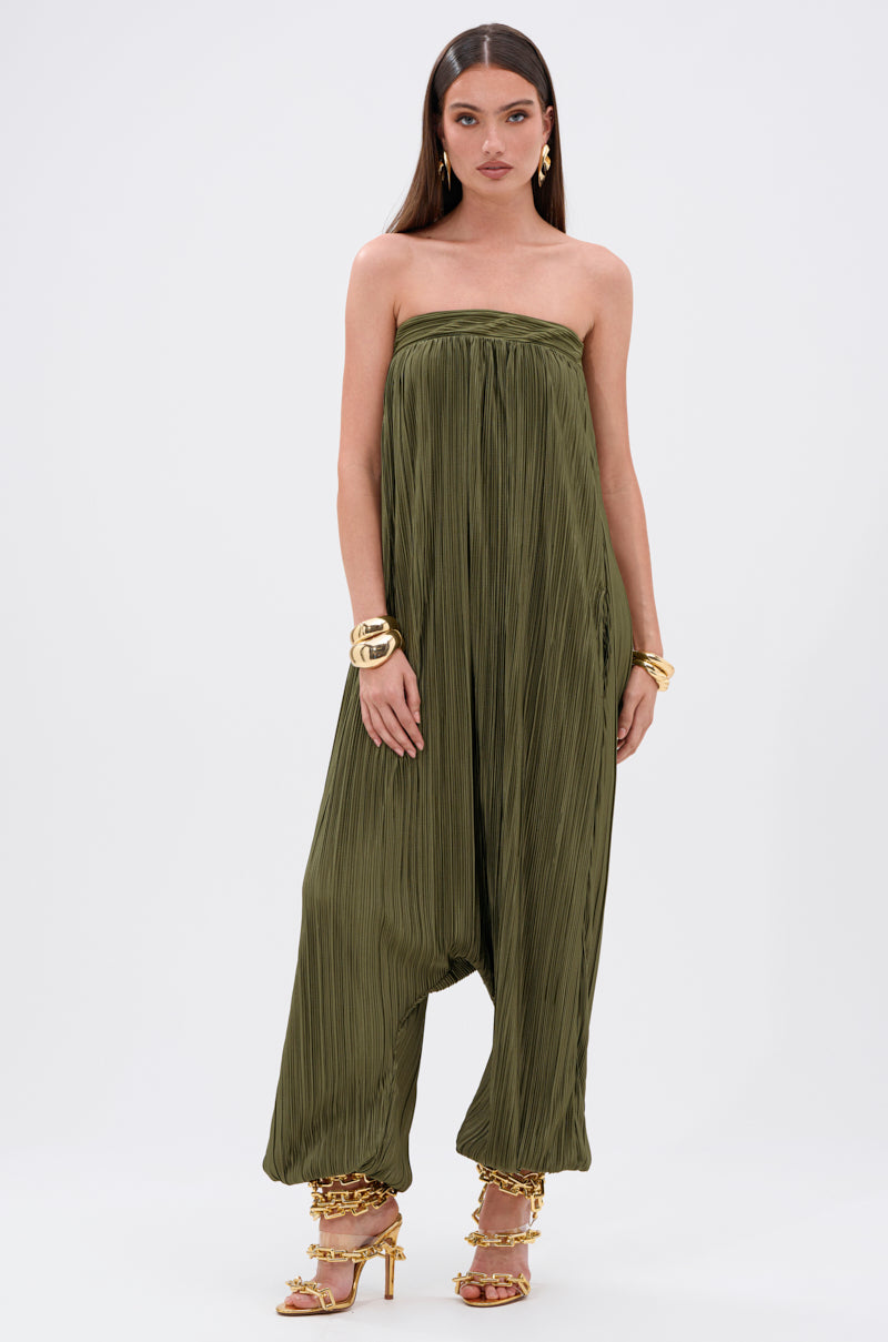 A woman wears the FROM THE CITY DROP CROTCH JUMPSUIT, a strapless olive pleated style with wide legs gathered at the ankles, styled with gold bracelets and strappy high-heeled sandals against a white background.
