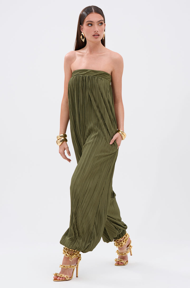 She wears the FROM THE CITY DROP CROTCH JUMPSUIT in olive satin—a strapless harem-style piece with loose, pleated legs cinched at the ankles—paired with gold hoop earrings, chunky bracelets, and strappy gold heels.