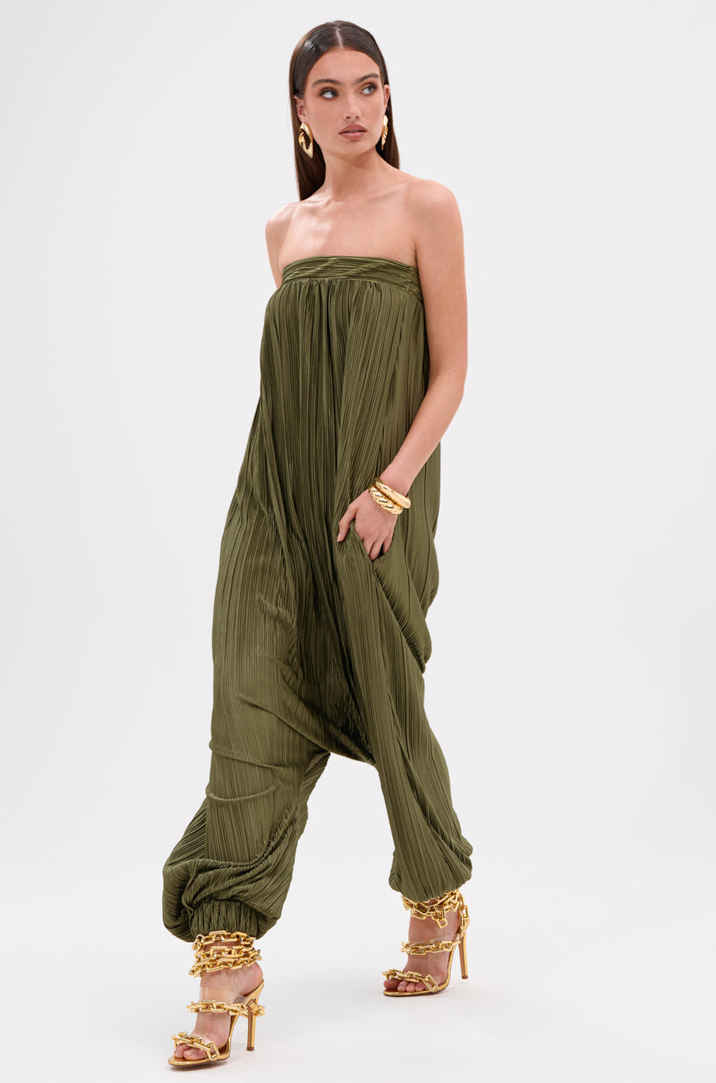 A woman models the FROM THE CITY DROP CROTCH JUMPSUIT—an olive green, silky satin, strapless jumpsuit with a pleated top and harem silhouette. She pairs it with gold hoop earrings, bracelets, and strappy sandals against a plain white background.