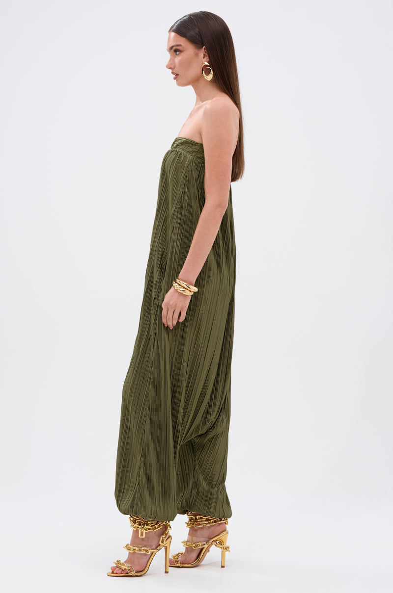 A woman in profile wears the FROM THE CITY DROP CROTCH JUMPSUIT in olive satin with a harem silhouette, gathered ankles, gold earrings, bracelets, gold high-heeled sandals, and long straight hair against a plain white background.