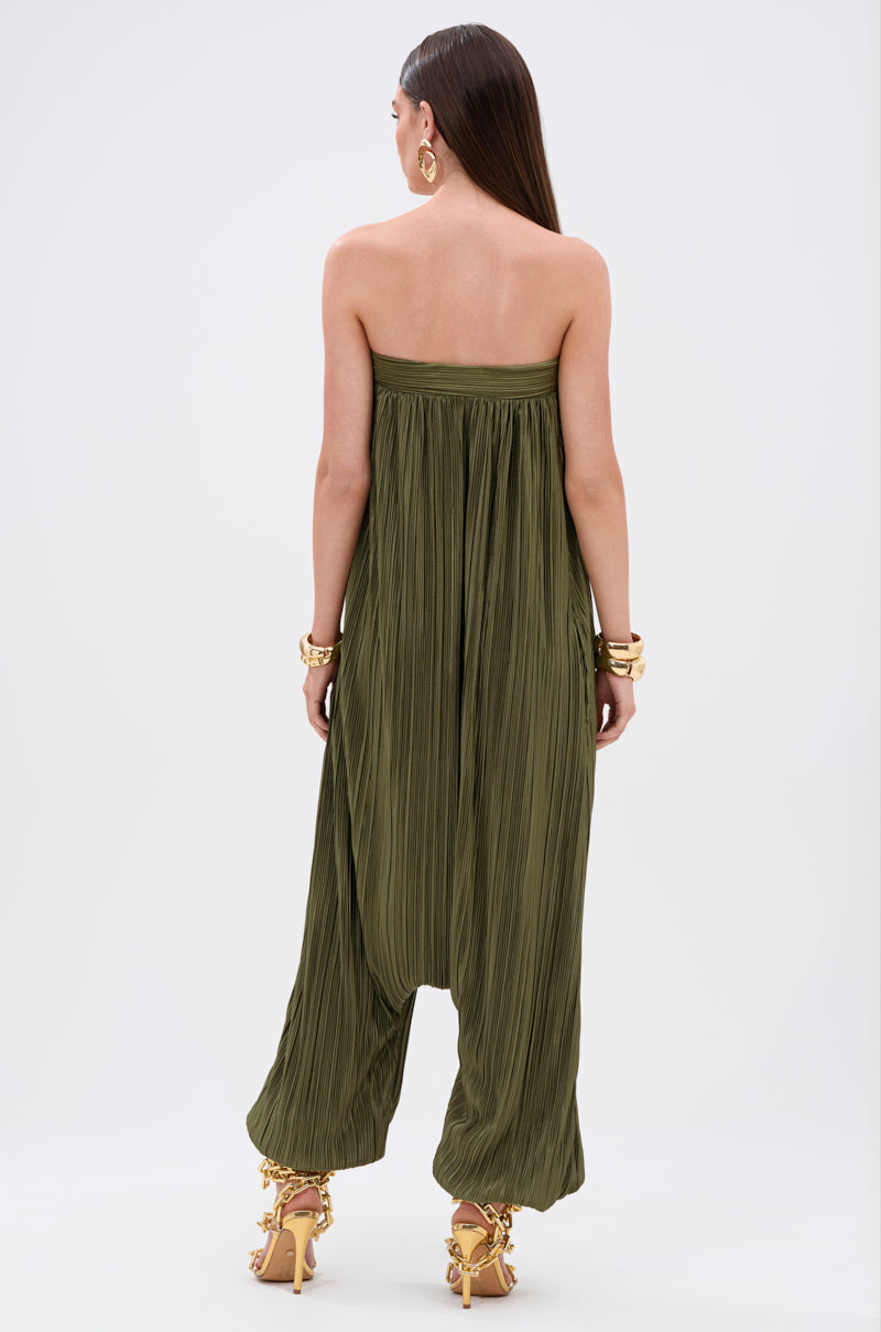 A woman faces away in the FROM THE CITY DROP CROTCH JUMPSUIT—an olive, strapless satin style with wide legs. She has long straight hair, gold earrings, bracelets, and high-heeled sandals against a white background.
