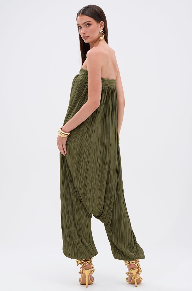 A woman with long straight hair, gold jewelry, and ornate high-heeled sandals poses slightly turned in the FROM THE CITY DROP CROTCH JUMPSUIT—an olive, strapless, pleated style—against a plain white background.