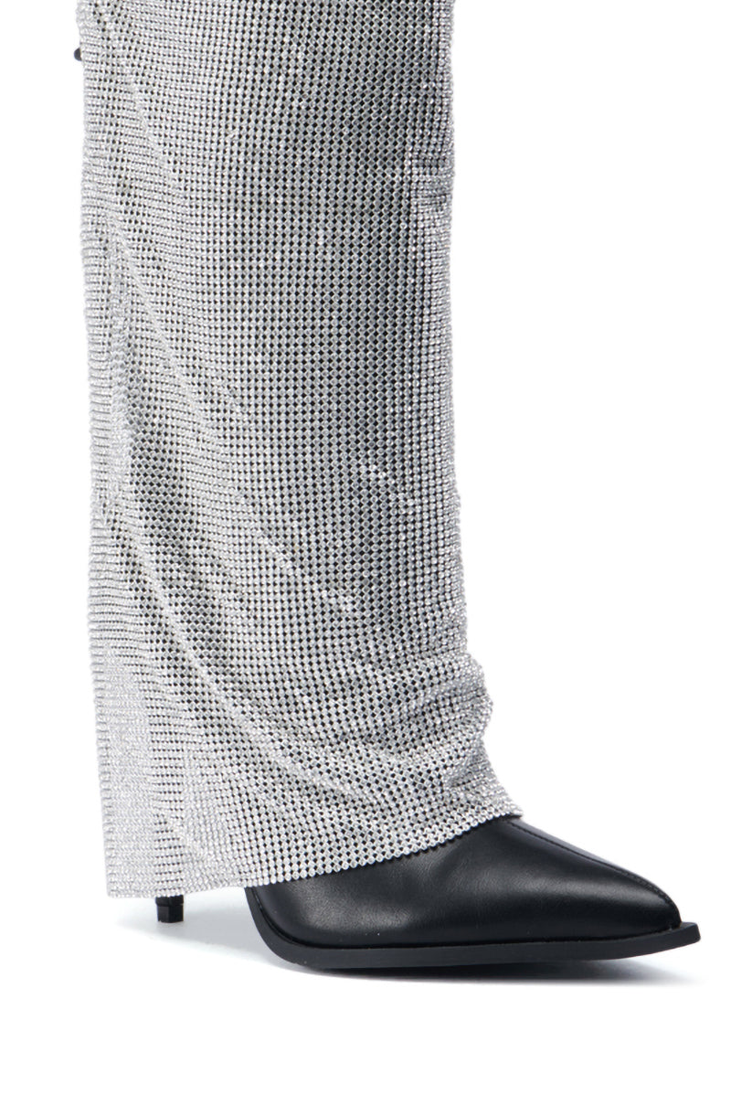 Close-up of a person's foot wearing the AZALEA WANG JACLYN EMBELLISHED STILETTO BOOT IN BLACK. The boot is partially covered by long, sparkling silver sequin pants, creating a glamorous and stylish look. The ensemble exudes elegance with its subtle rhinestone mesh overlay accents.