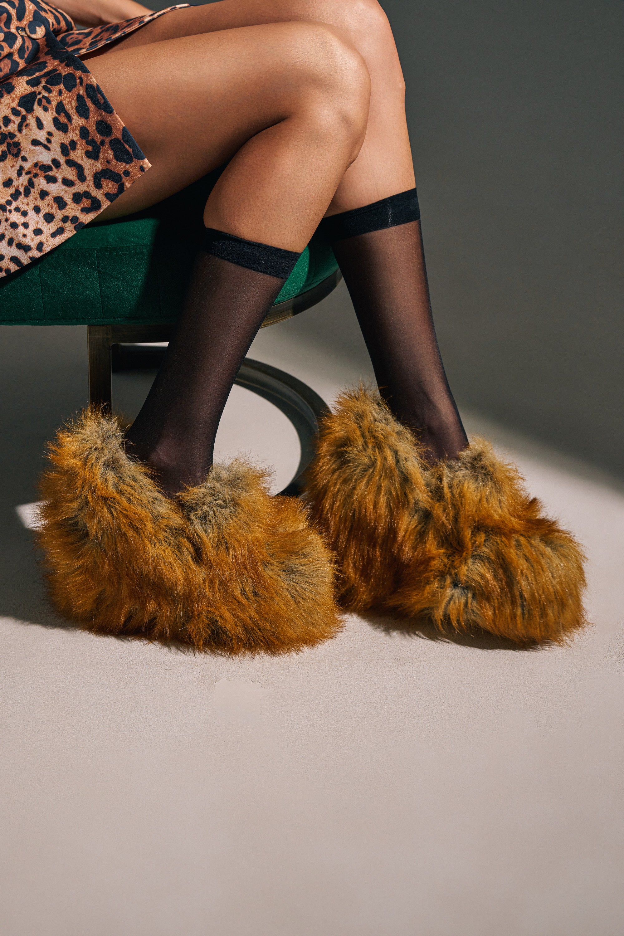 A person sits cross-legged on a green chair wearing sheer black knee-high socks, a leopard print skirt, and AZALEA WANG JAEKE FOX FUR WEDGE PUMP IN BROWN with comfort cushion insoles.