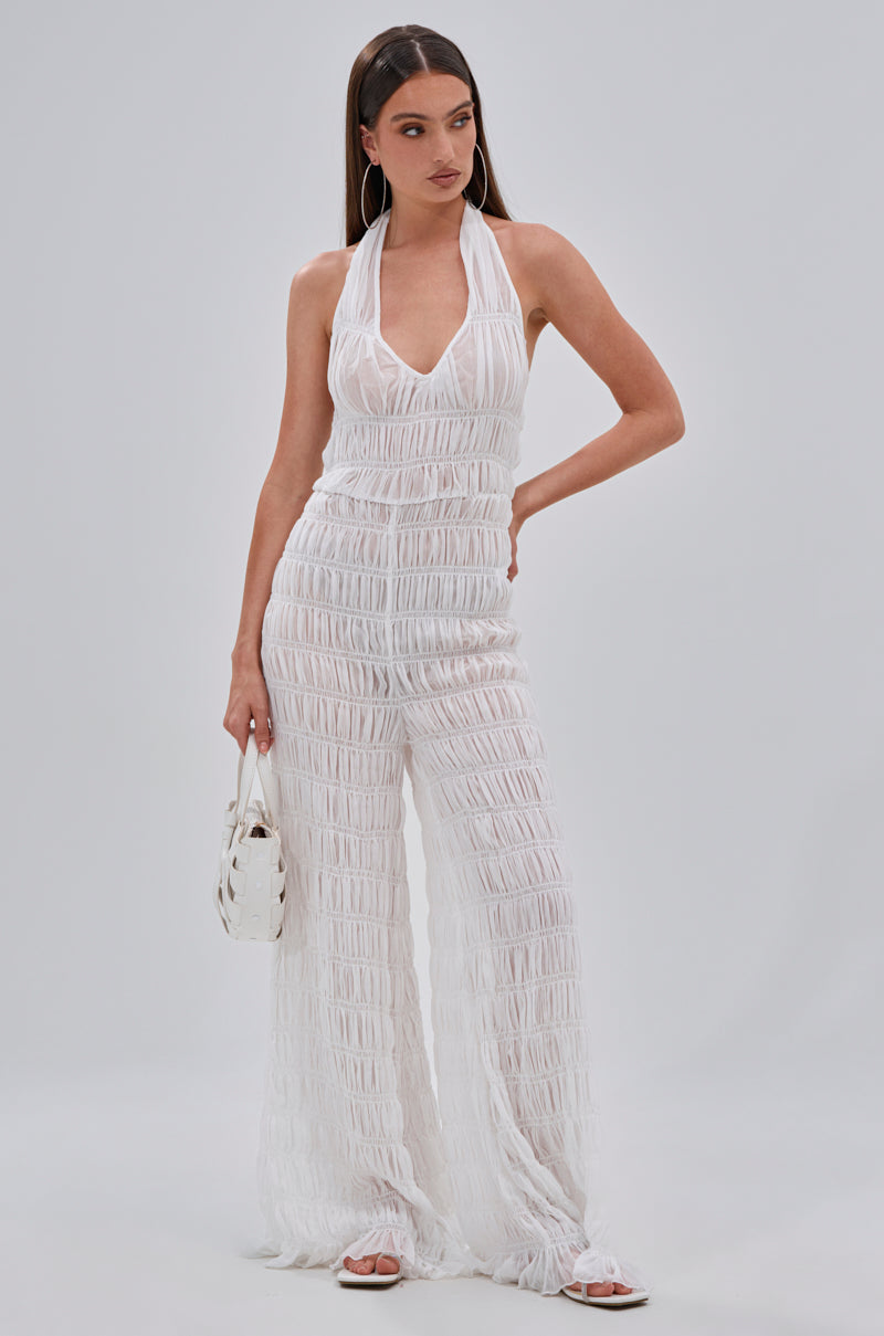 A woman poses in the SUMMERTIME SHINE MESH JUMPSUIT with wide legs and a halter neck, paired with sandals, a white handbag, and hoop earrings. Her long brown hair is straight—ideal for vacation glam.