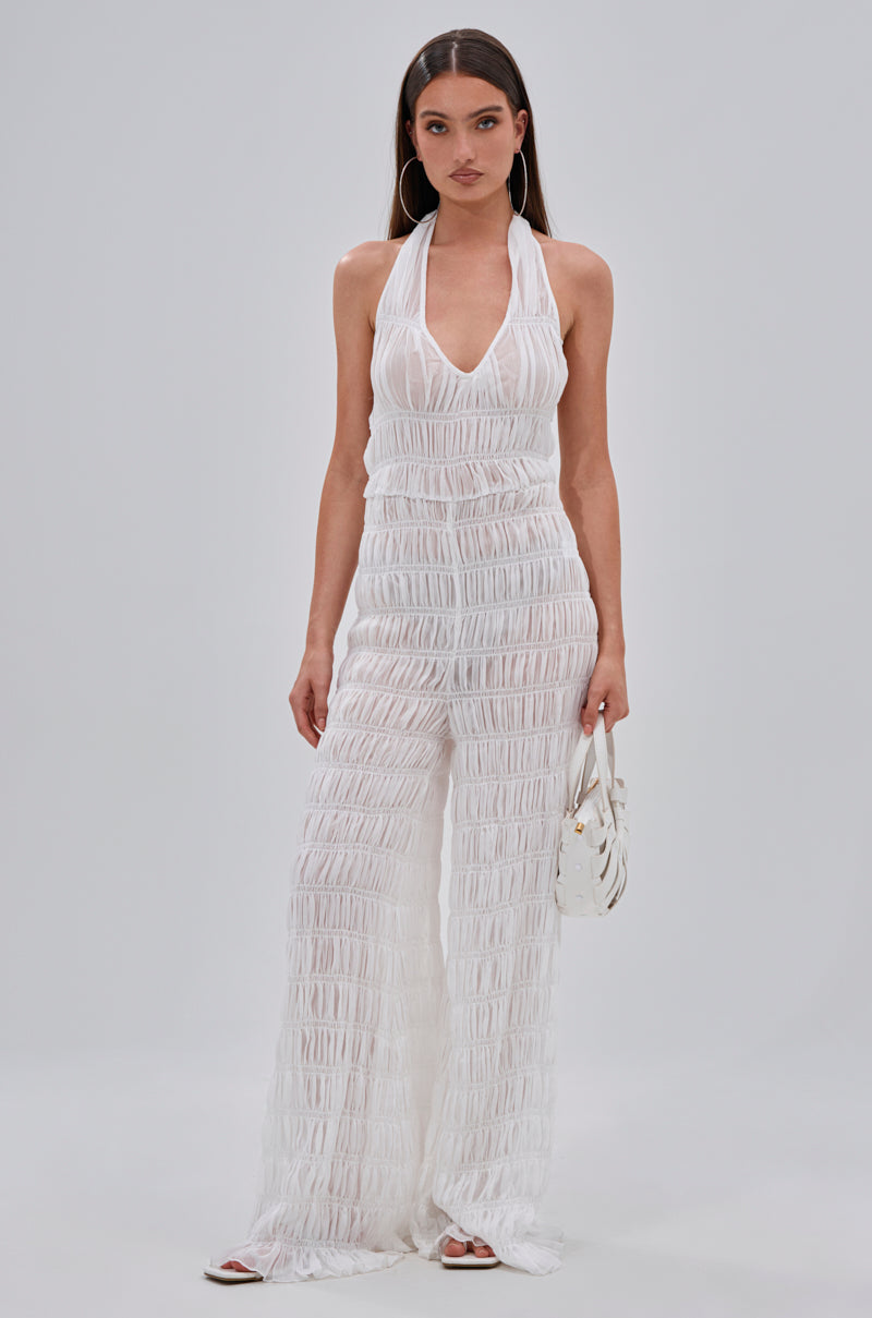 A woman poses against a plain backdrop in the SUMMERTIME SHINE MESH JUMPSUIT—a sleeveless, deep V-neck white sheer piece ideal for beach parties or vacations. She accessorizes with a small white handbag, hoop earrings, and open-toe shoes.