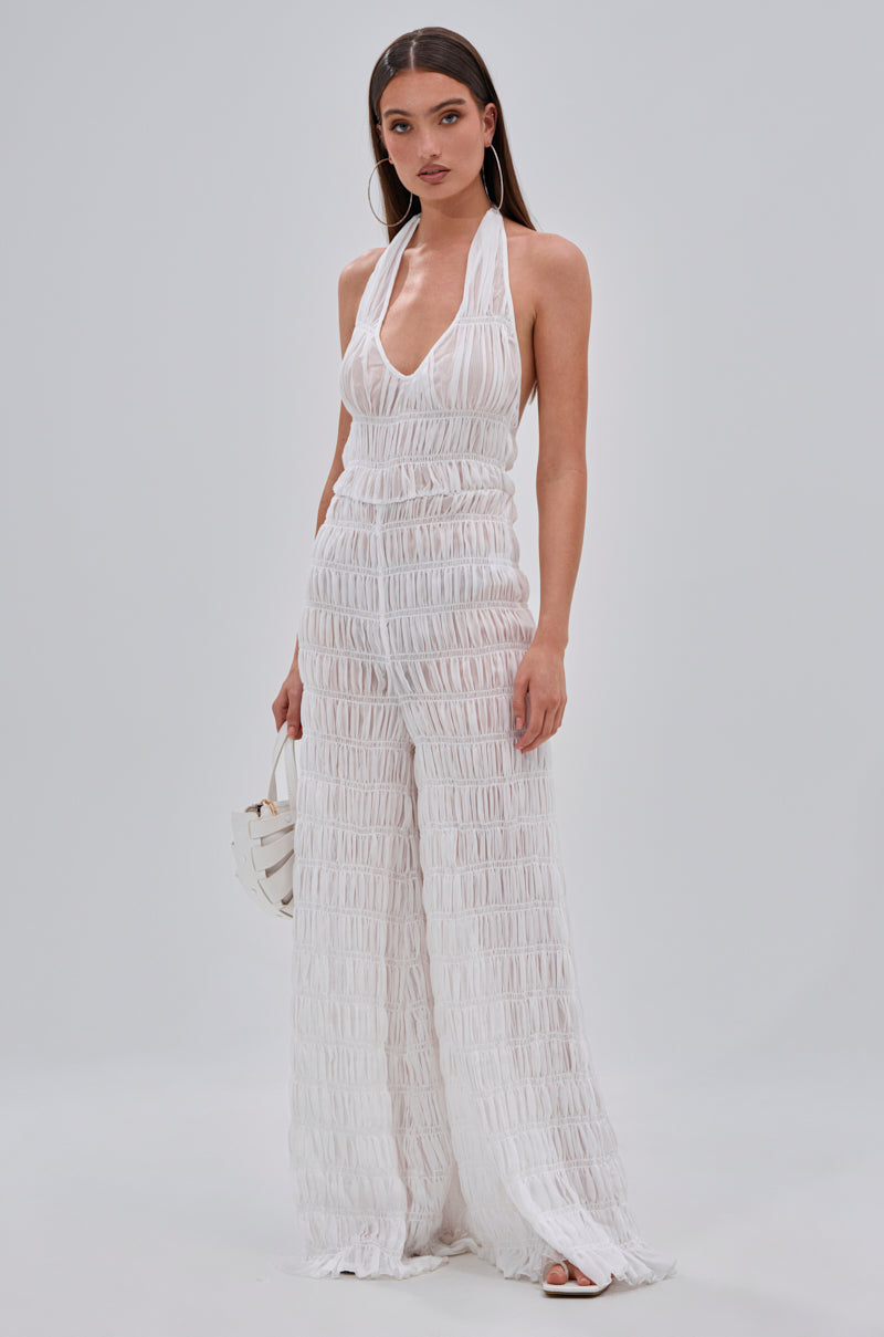 A woman stands confidently against a plain backdrop in the SUMMERTIME SHINE MESH JUMPSUIT, featuring a textured, pleated design. She accessorizes with a small white handbag—perfect style for beach parties or vacation glam.