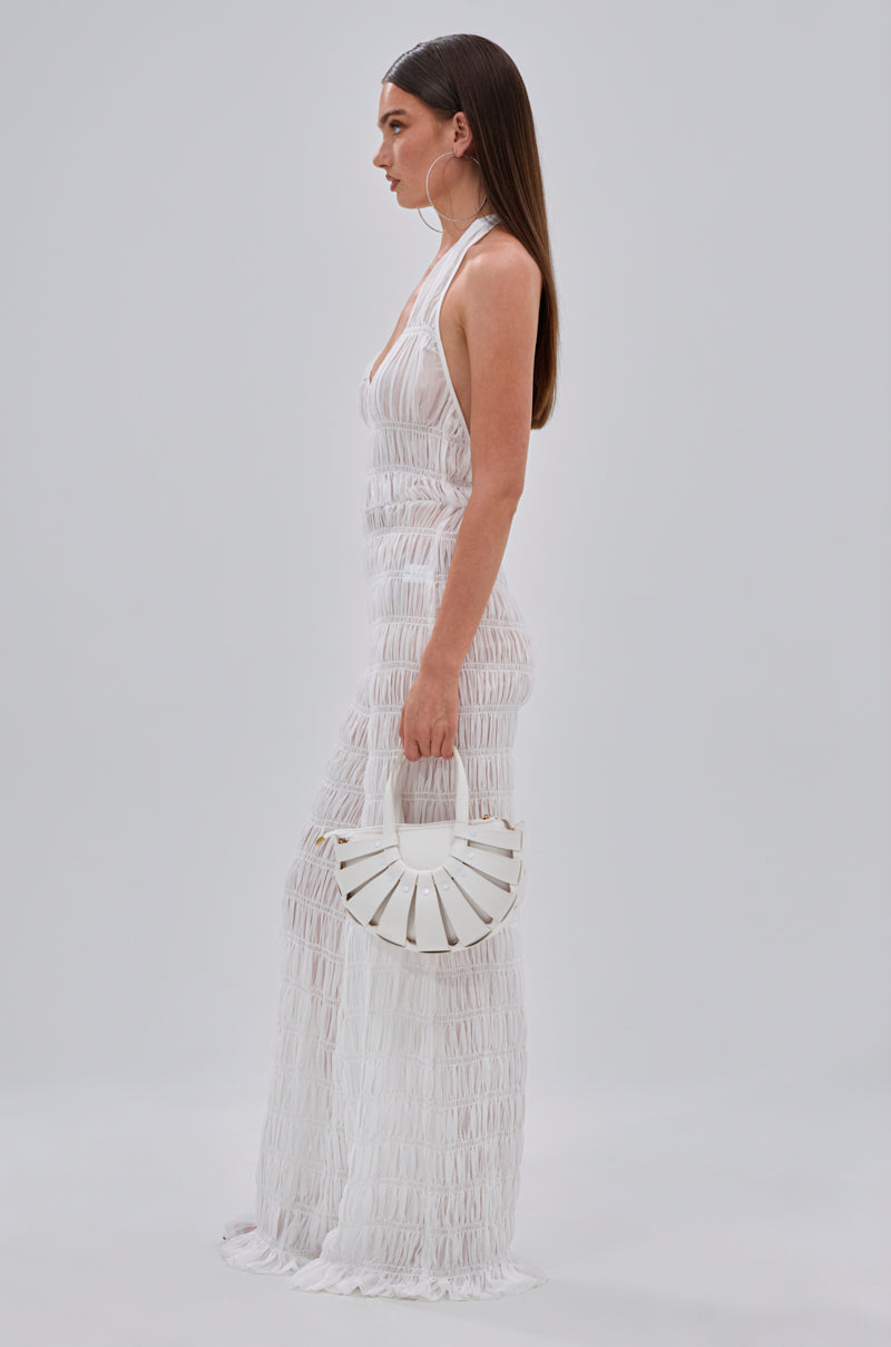A woman with long brown hair stands in profile against a light gray background, wearing the SUMMERTIME SHINE MESH JUMPSUIT and holding a white semi-circular handbag—her look is perfect for beach parties and vacation glam.
