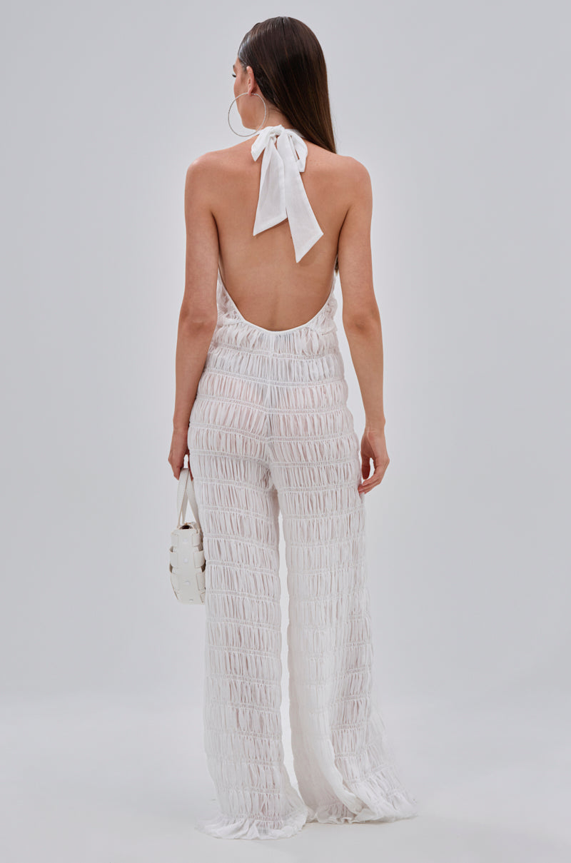 A woman, back turned, wears the SUMMERTIME SHINE MESH JUMPSUIT—a white, backless halter jumpsuit with ruched mesh—ideal for vacation glam. She holds a small white bag; her long hair stands out against the light background.