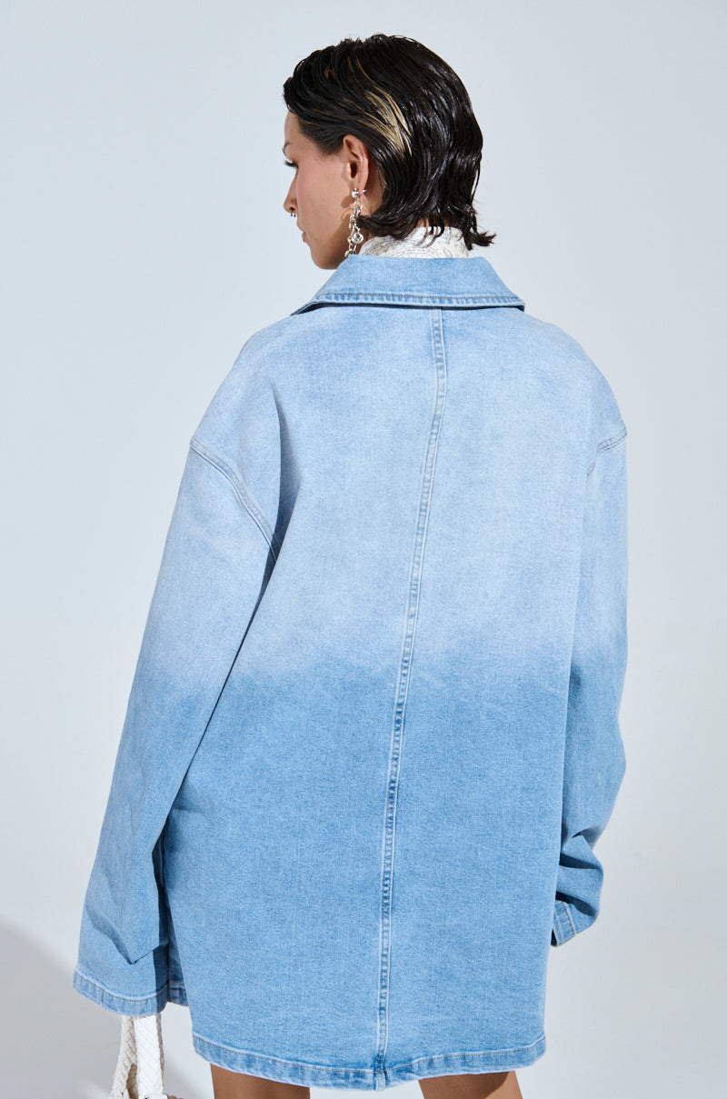 A person with short slicked-back hair, showcasing a casual-cute style in a COOL GIRL OVERSIZED OMBRE DENIM SHIRT DRESS, holds a white textured bag against a plain, light background.