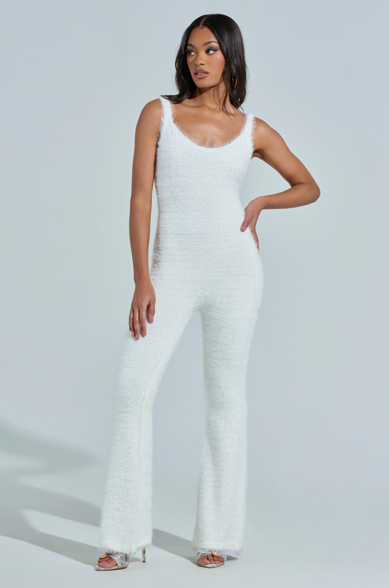 A woman poses confidently against a plain background in the STAYING IN FUZZY KNIT JUMPSUIT with flared legs, her long hair flowing. One hand is on her hip while the other rests by her side, and she completes the chic loungewear look with stylish open-toed shoes.