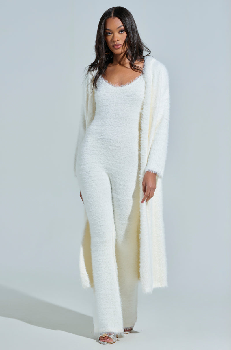 Draped in the "Staying In Fuzzy Knit Jumpsuit" with a matching cardigan, a woman epitomizes chic loungewear. She completes the cozy look with sandals, elegantly standing against a light background.