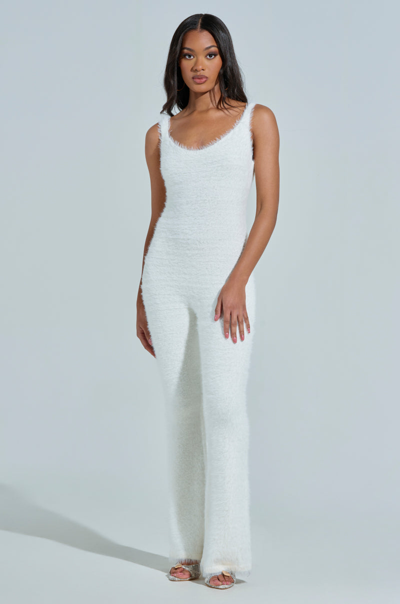 A woman wearing the STAYING IN FUZZY KNIT JUMPSUIT, in white and sleeveless, stands against a light background. Her long dark hair cascades freely as she wears open-toed shoes. With a neutral expression and one hand by her side, she exudes an elegant loungewear chic vibe.