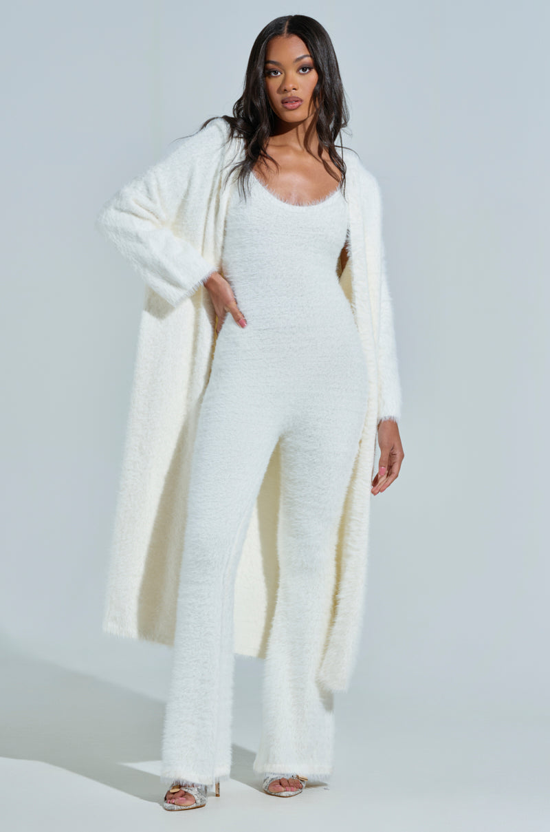 Against a plain background, a woman stands confidently in the STAYING IN FUZZY KNIT JUMPSUIT featuring elegant flared pants. She complements the outfit with a matching faux mohair coat, long hair, and clear heels.