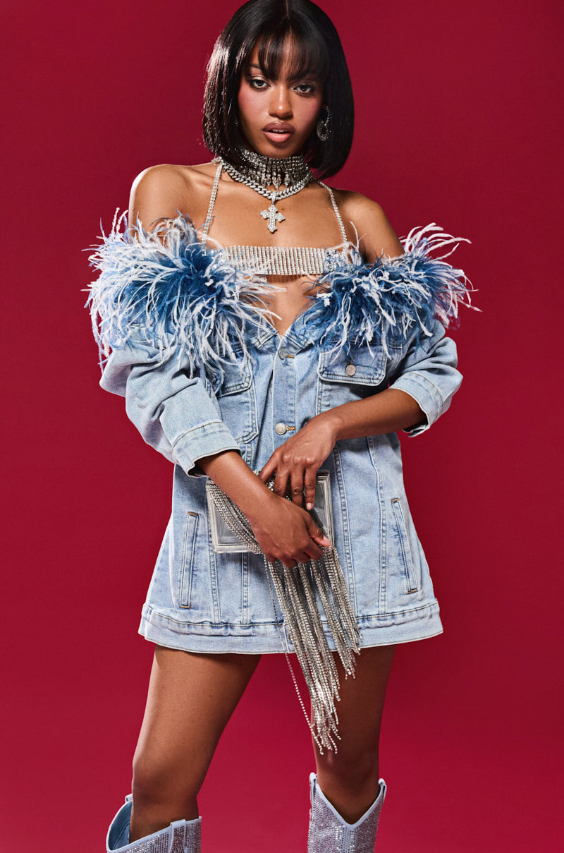 A person stands against a red background wearing the CHIC AF DENIM MINI DRESS WITH FEATHERS, paired with a glittery top, rhinestone accessories, a sparkly clutch with fringe, and tall shiny boots, showcasing an off-the-shoulder design with flair.