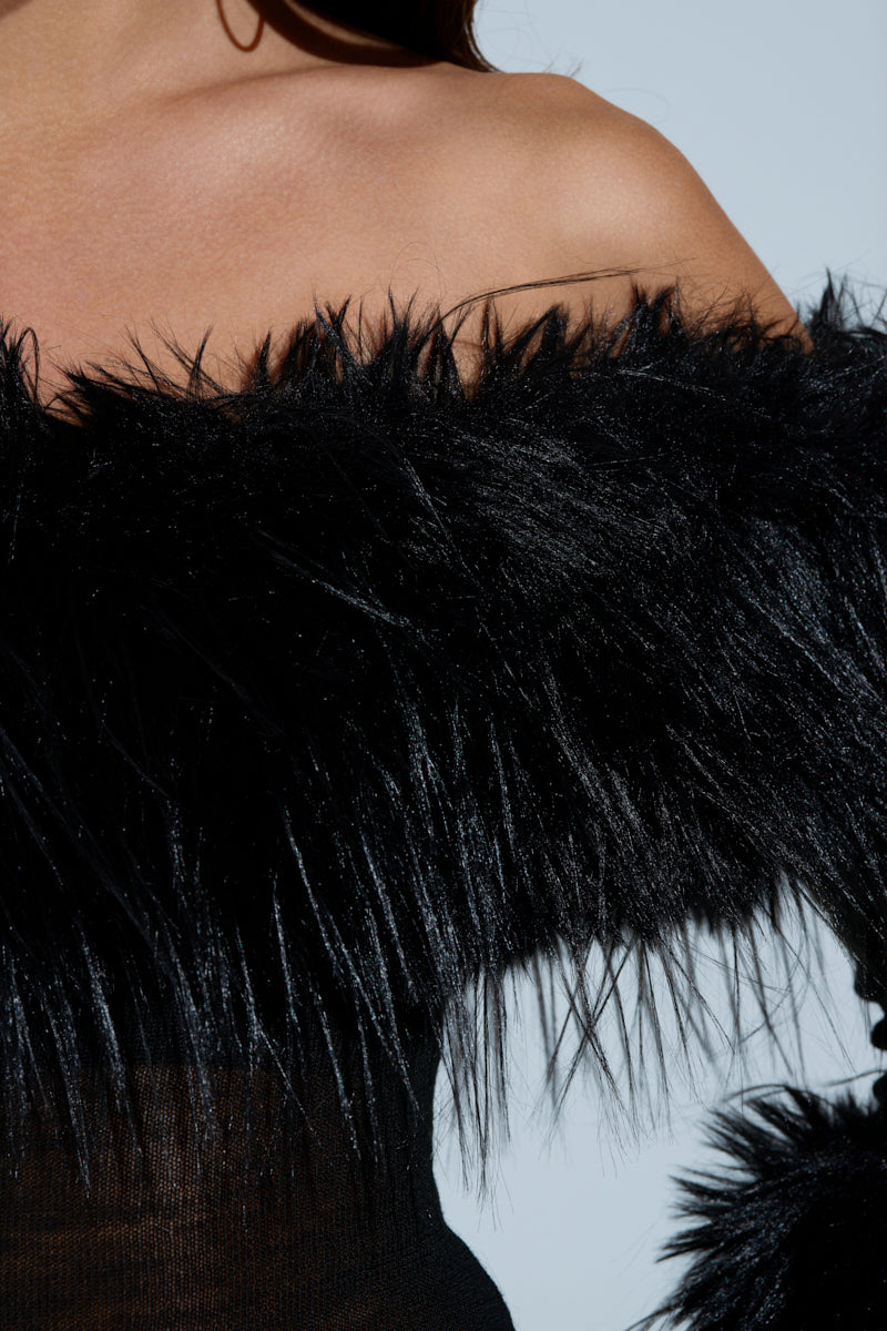 A close-up features a person in the STAR OF THE SHOW FUR MINI DRESS, a black, fuzzy off-shoulder garment with a strapless neckline. The luxurious fabric drapes over the shoulders and upper arms against a plain, pale background.