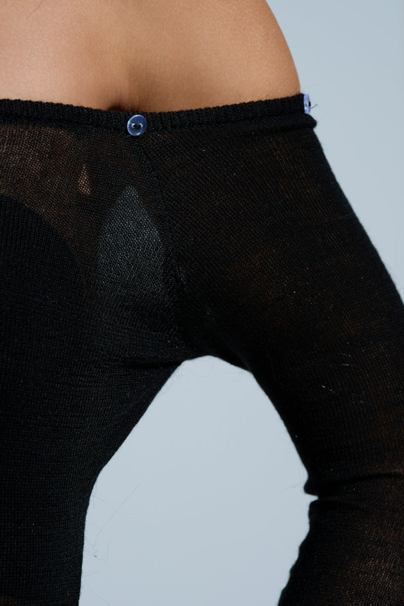 Close-up of a person in a mini dress named "Star of the Show," highlighted by its sheer knit black fabric and an off-shoulder design, fastened with a small blue button at the shoulder against a soft light blue gradient background.