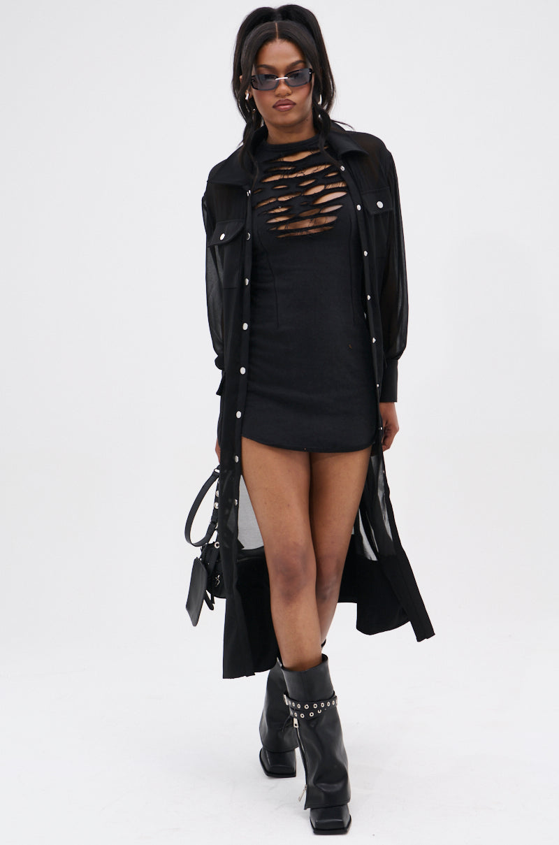 Wearing sunglasses, a long sheer black shirt, black studded boots, and carrying a small black handbag, a woman walks confidently in the DIXIE SHREDDED MINI DRESS—a curve-hugging mini with shredded details—against a plain background.