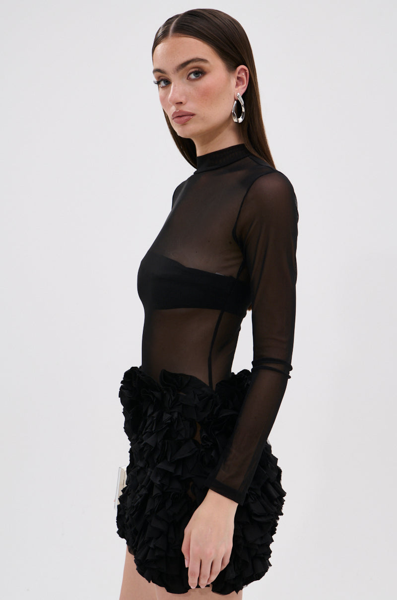 A woman with long straight brown hair models the KELYN MESH MINI DRESS IN BLACK, featuring sheer long sleeves and textured ruffle detailing, against a plain white background. She wears hoop earrings and neutral makeup.