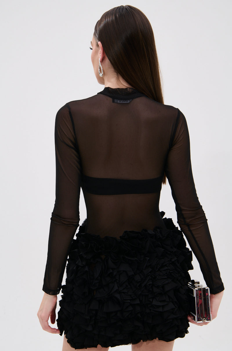 A woman with straight brown hair wears the KELYN MESH MINI DRESS IN BLACK, exuding sheer sophistication. Facing away, she holds a small rectangular clutch bag in her left hand.