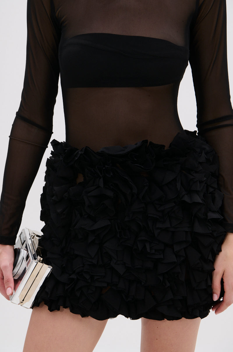 Wearing the KELYN MESH MINI DRESS IN BLACK, which features sheer black mesh and textured ruffle detailing on the skirt, a person exudes sophistication while holding a metallic clutch. The image is cropped from shoulders to mid-thigh.