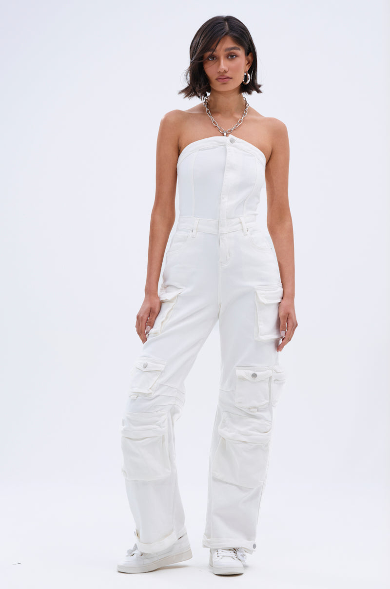 A woman with short dark hair and a neutral expression wears the LINDSAY DENIM CARGO JUMPSUIT IN WHITE, featuring cargo pockets. Styled with white sneakers and a silver chain necklace, she stands against a plain white background.