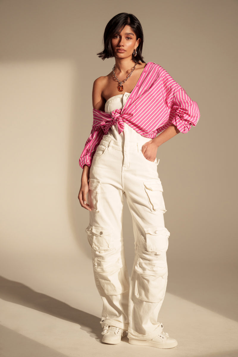 A woman stands confidently in the LINDSAY DENIM CARGO JUMPSUIT IN WHITE with sneakers, a strapless top, and a pink striped button-up shirt tied at the waist—showcasing utility fashion with her dark hair in a side part.
