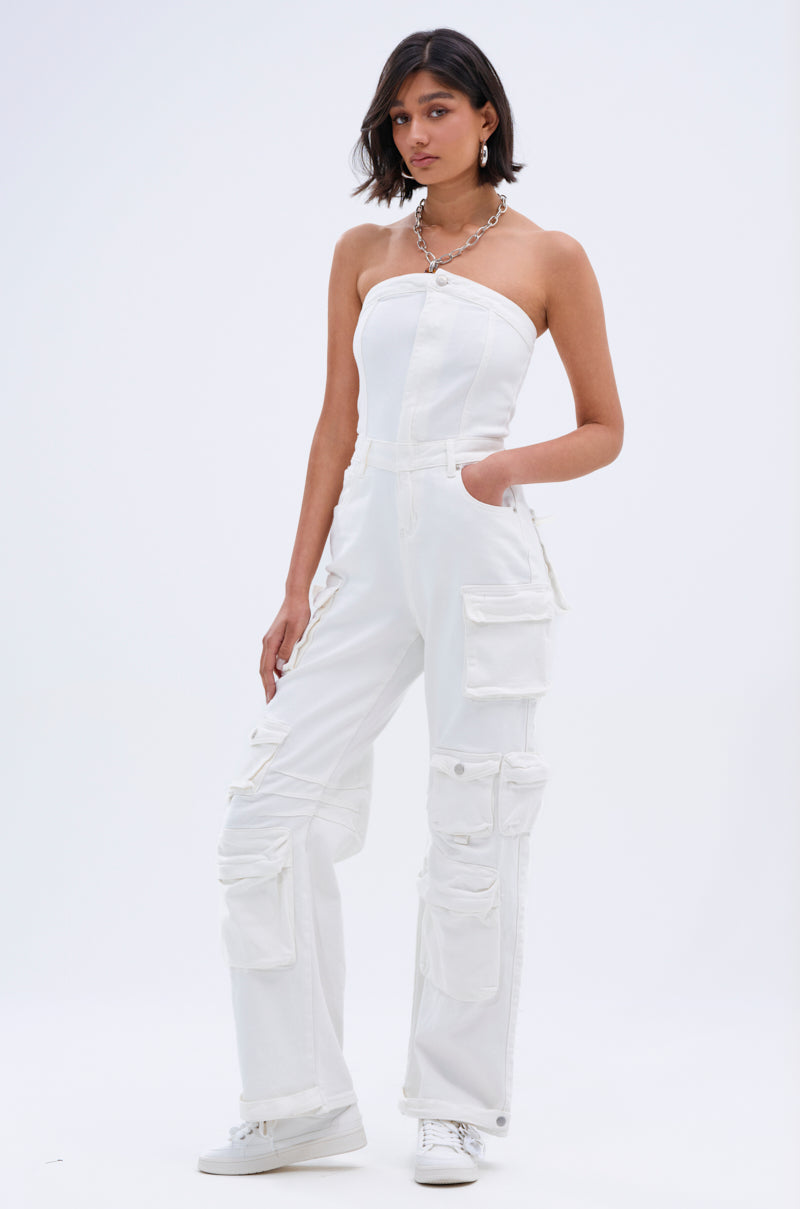 A woman with short dark hair poses against a plain background in the LINDSAY DENIM CARGO JUMPSUIT IN WHITE, styled with earrings, a chain necklace, white sneakers, and one hand in her pocket for a utilitarian look.